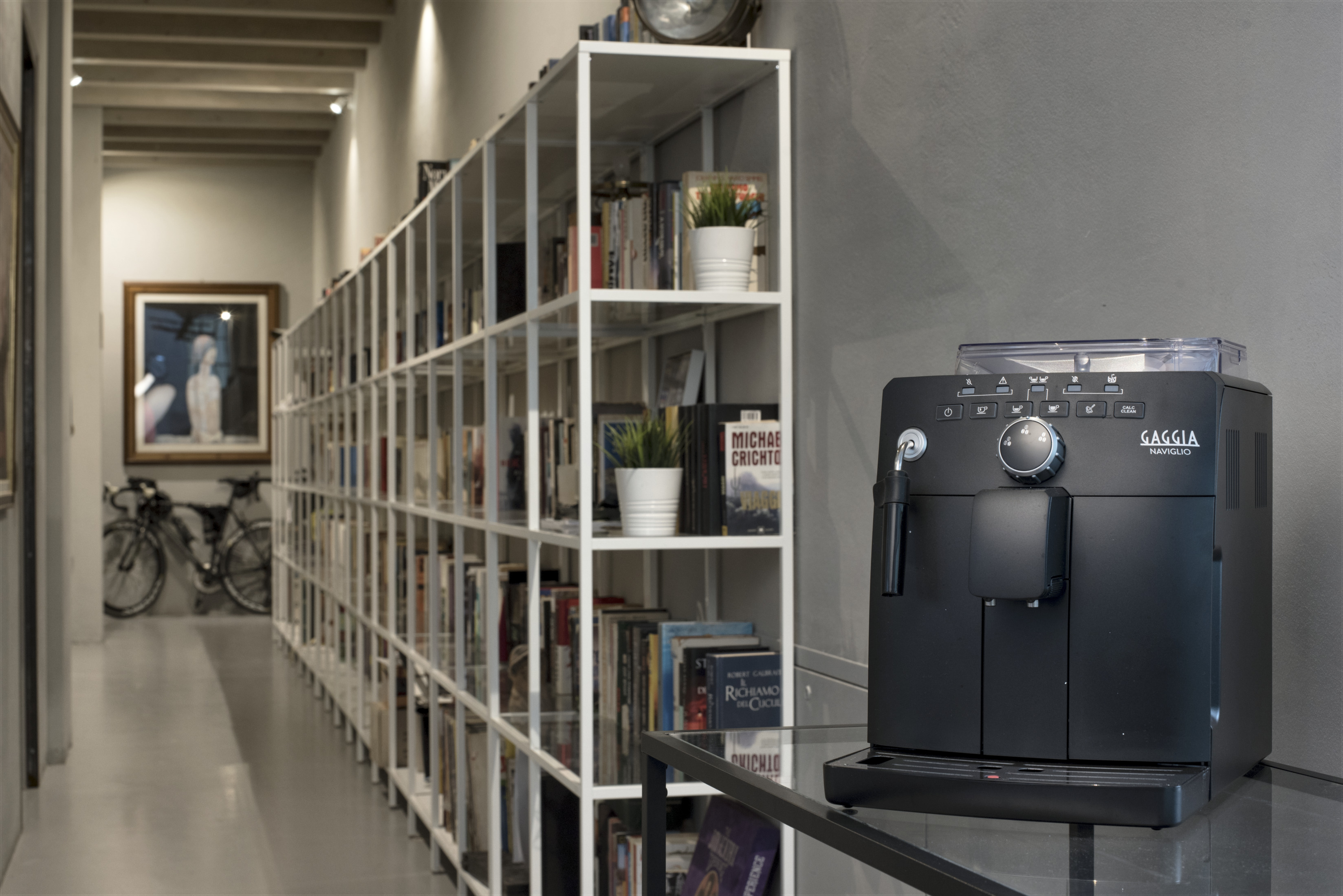Czarny ekspres do kawy stoi na szklanym stole obok regału z książkami.
