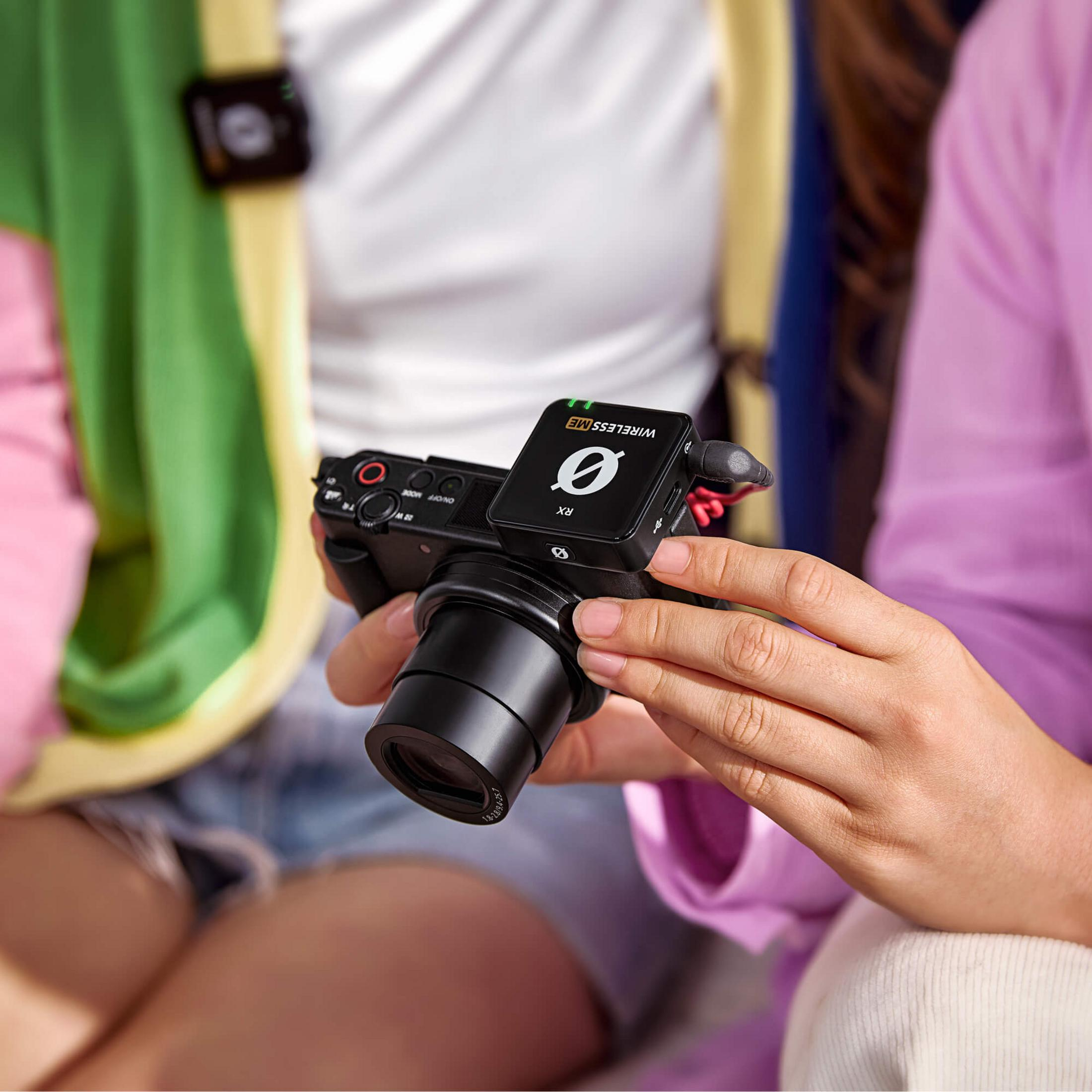 Osoba trzyma aparat z mikrofonem. Zielone i fioletowe ubrania w tle.