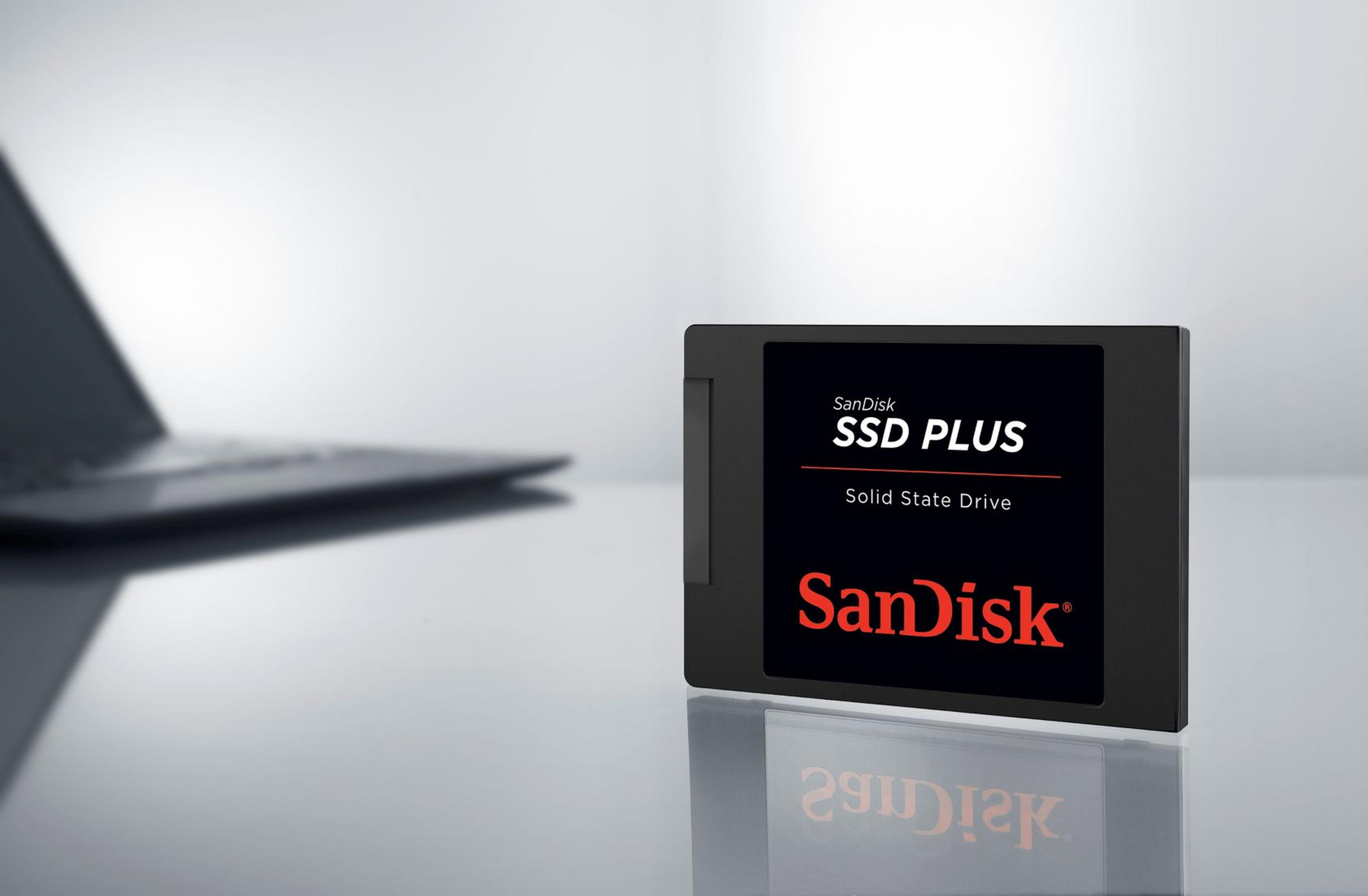SanDisk SSD PLUS Solid State Drive na czarnej powierzchni z laptopem w tle.