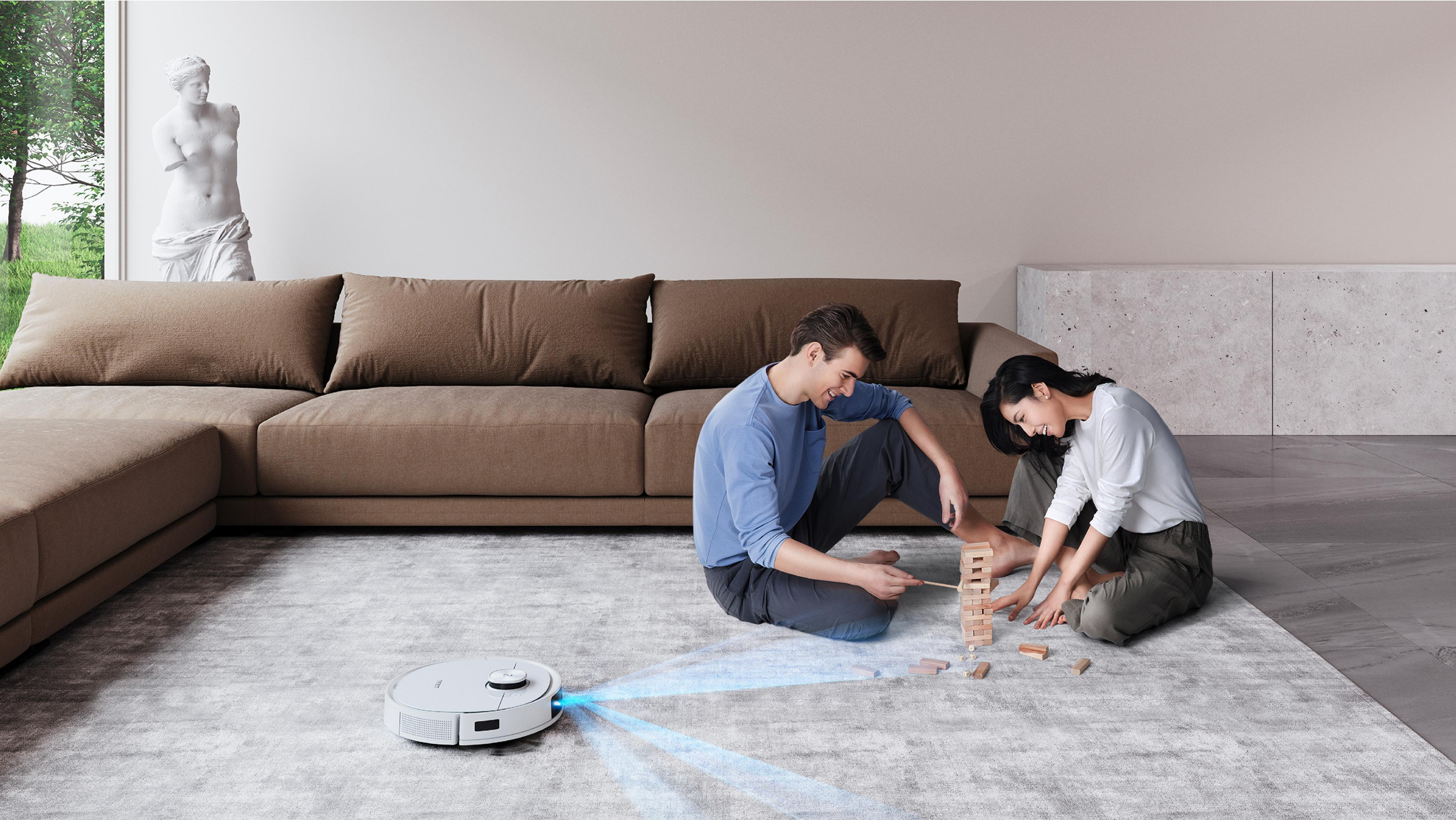Un robot aspirateur nettoie. Un couple joue au Jenga sur un tapis devant un canapé.