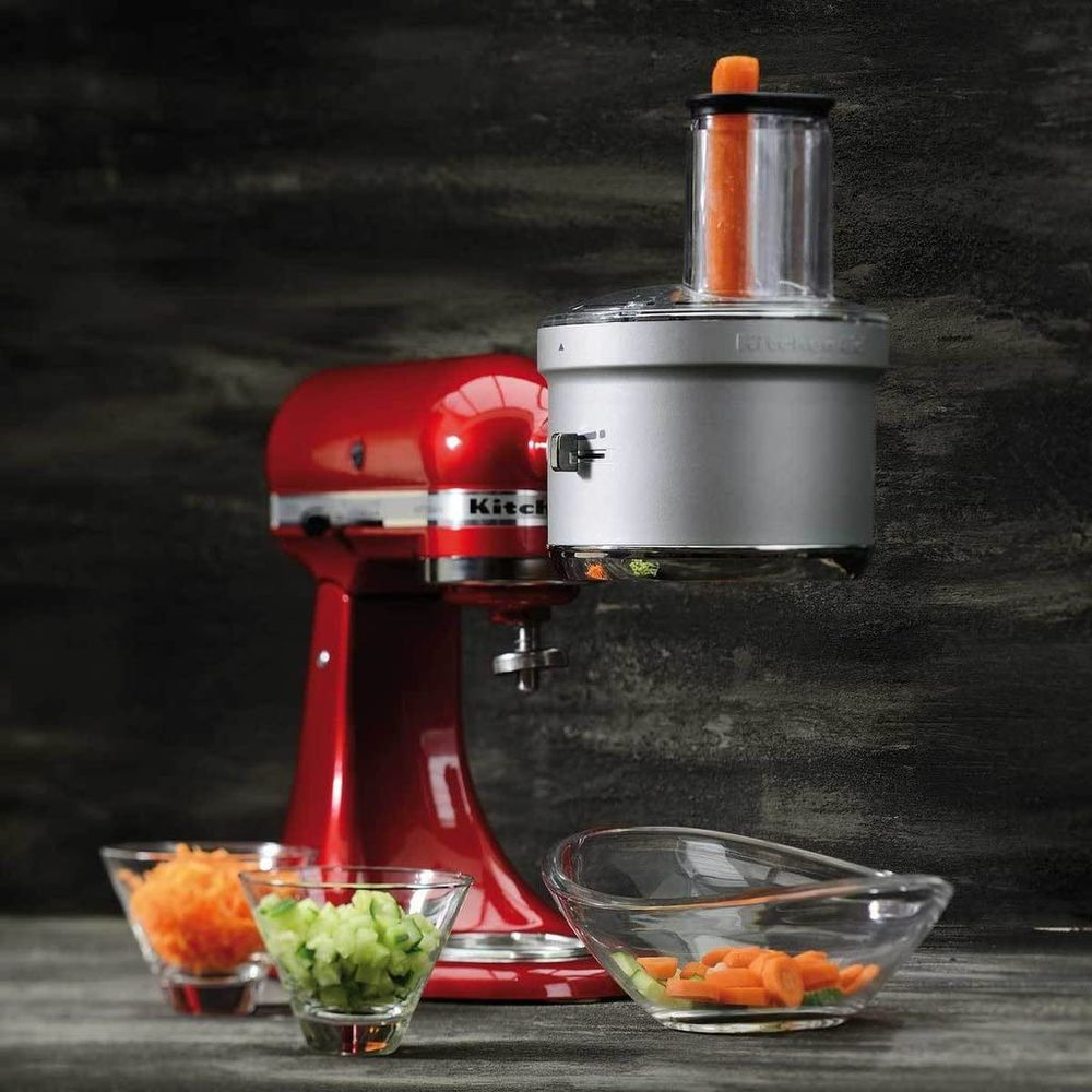 Un robot da cucina KitchenAid rosso con accessorio argento che processa carote. Vicino ci sono ciotole con carote grattugiate e cetrioli a cubetti.