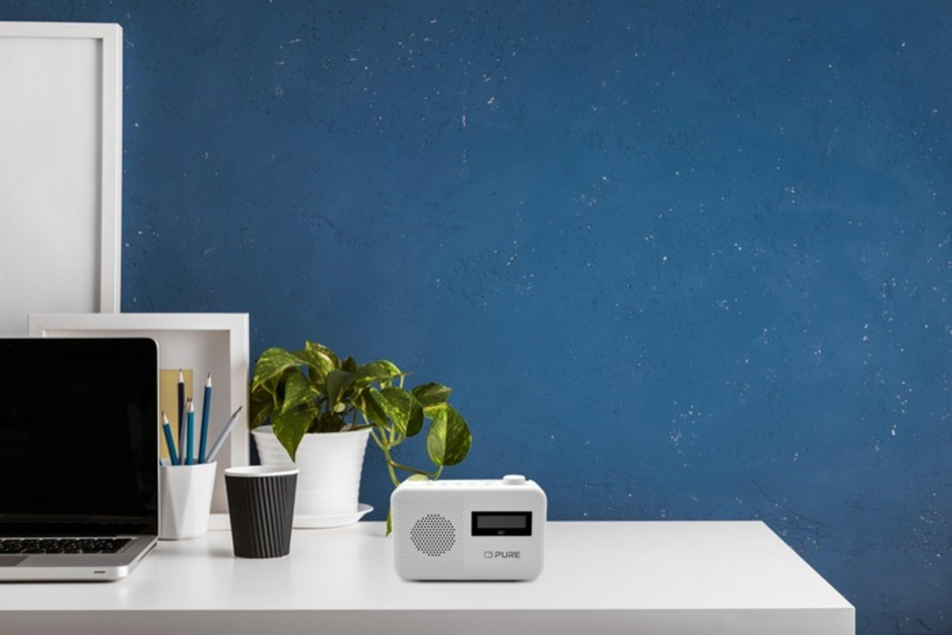 Białe radio stoi na biurku obok laptopa, rośliny i kubka.