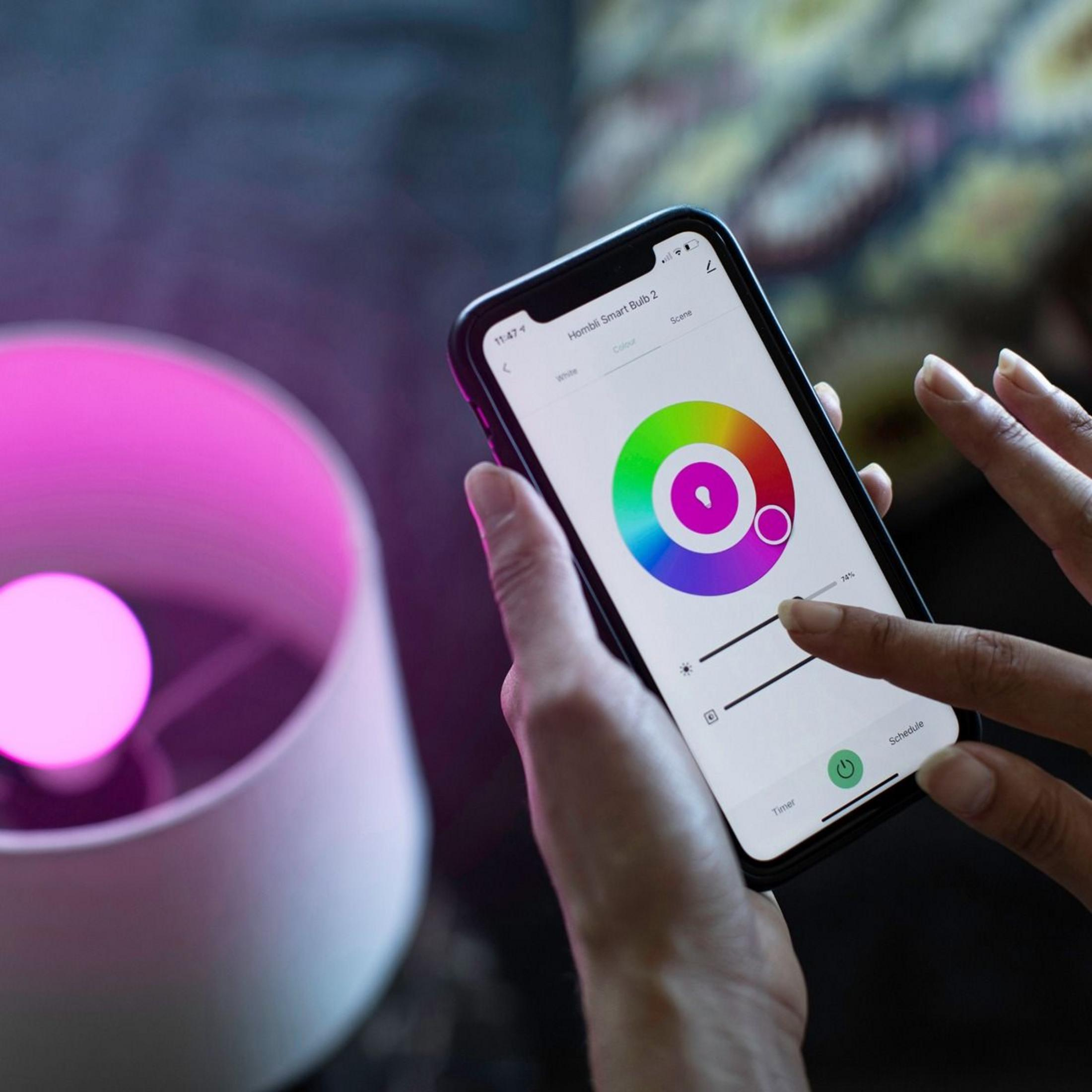 Dłoń trzyma smartfon z aplikacją koła kolorów, obok różowej żarówki smart.