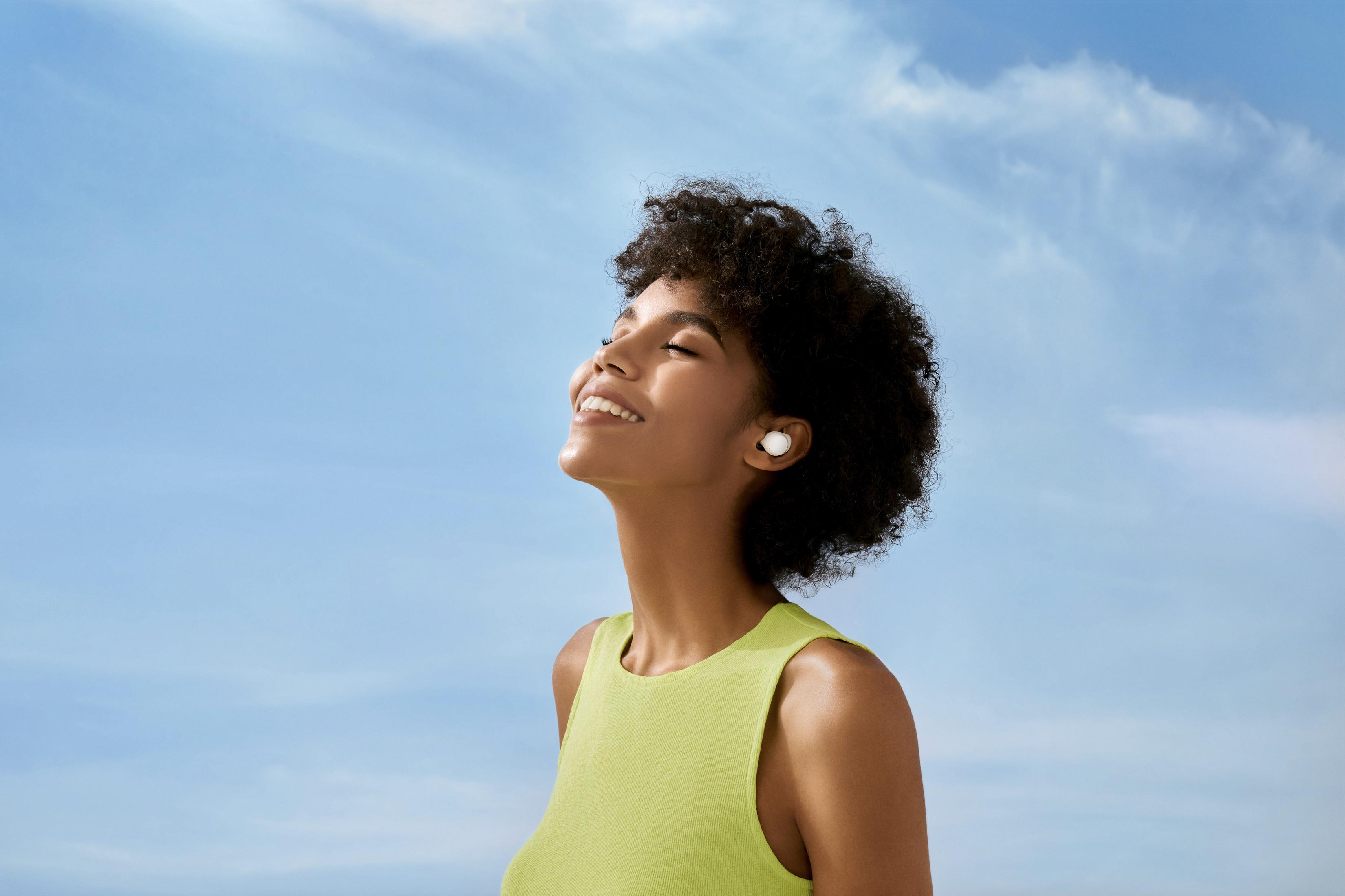 Kobieta z kręconymi włosami uśmiecha się z zamkniętymi oczami, nosi słuchawki.