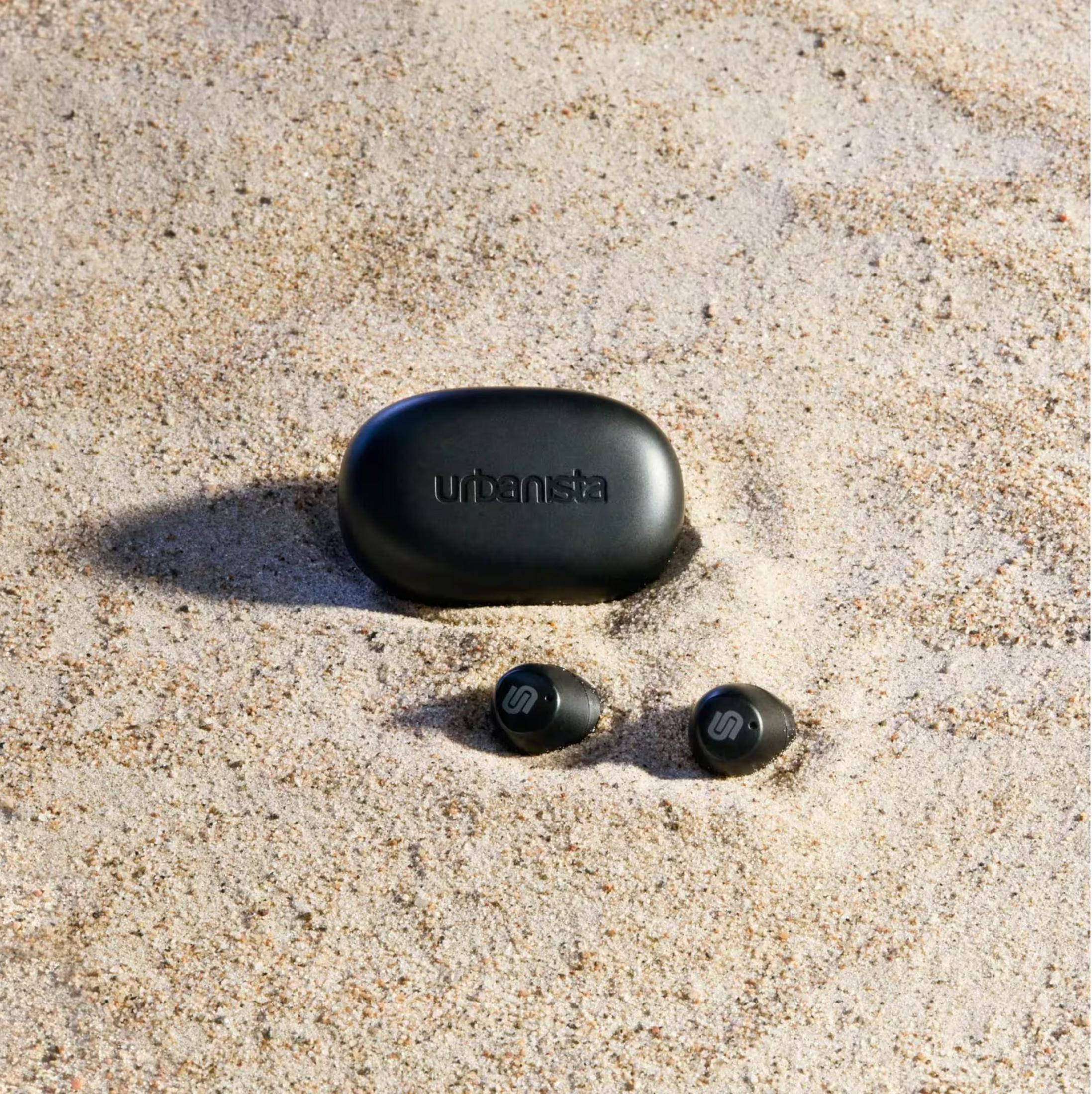 Zwarte oordopjes en oplaadcase in het zand.