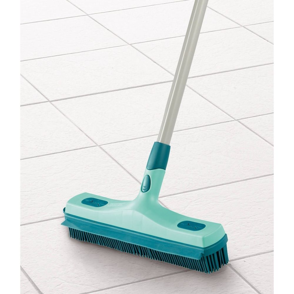 Una scopa verde e turchese con setole su un pavimento piastrellato bianco.