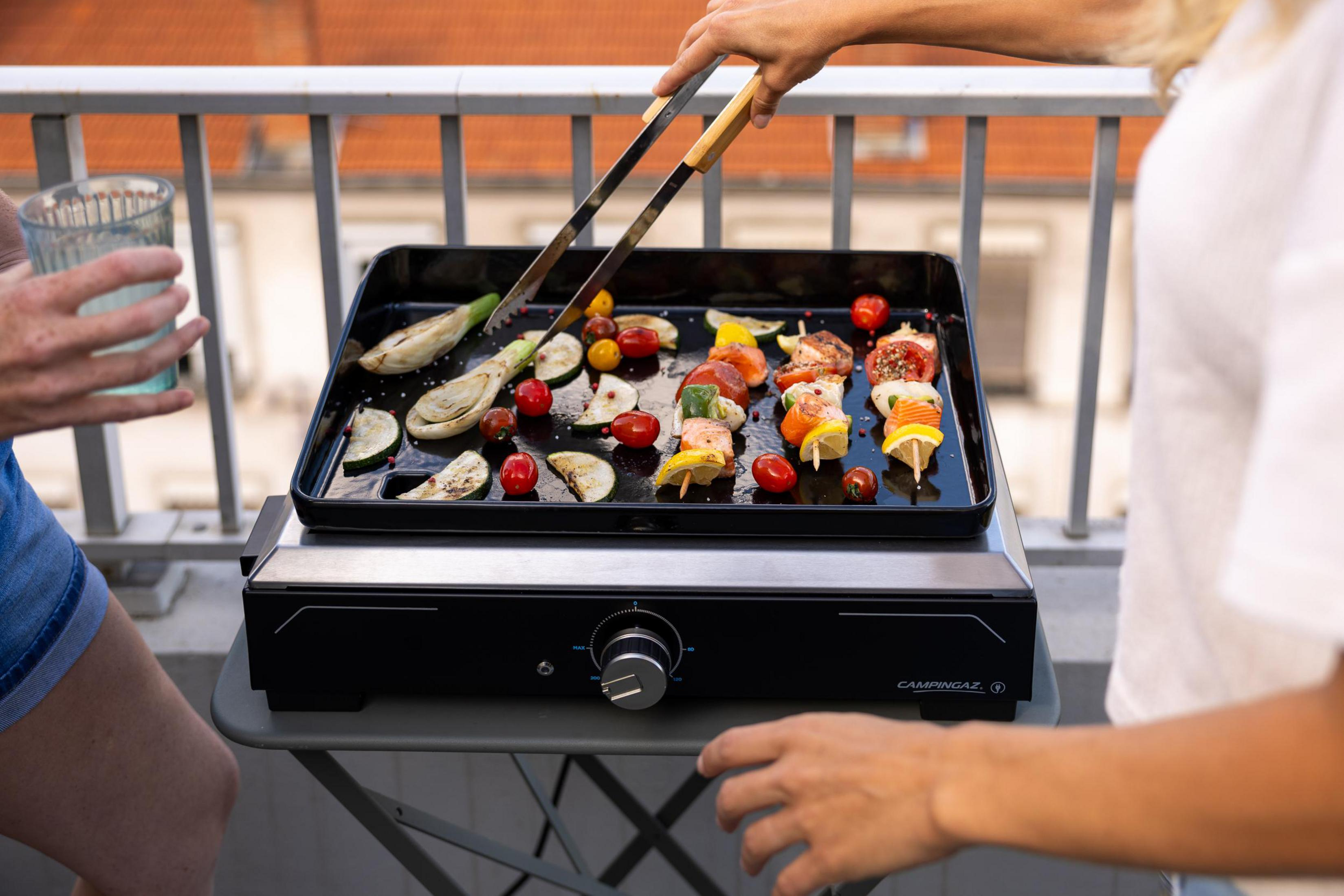 Zwei Personen grillen Spieße mit Lachs und Gemüse auf einem Balkon.