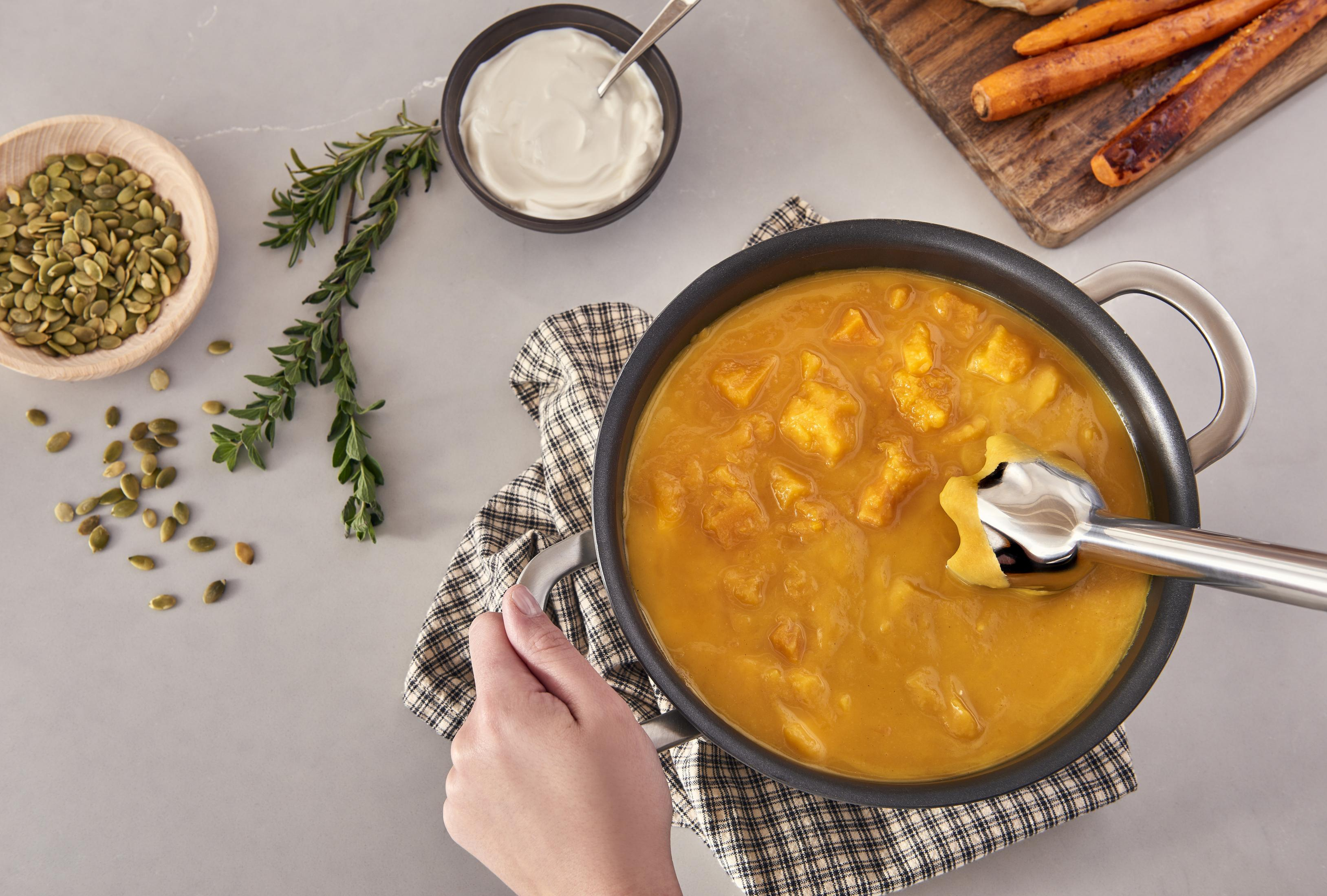 Ręka używa blendera do zupy do puree z pomarańczowej zupy w garnku.