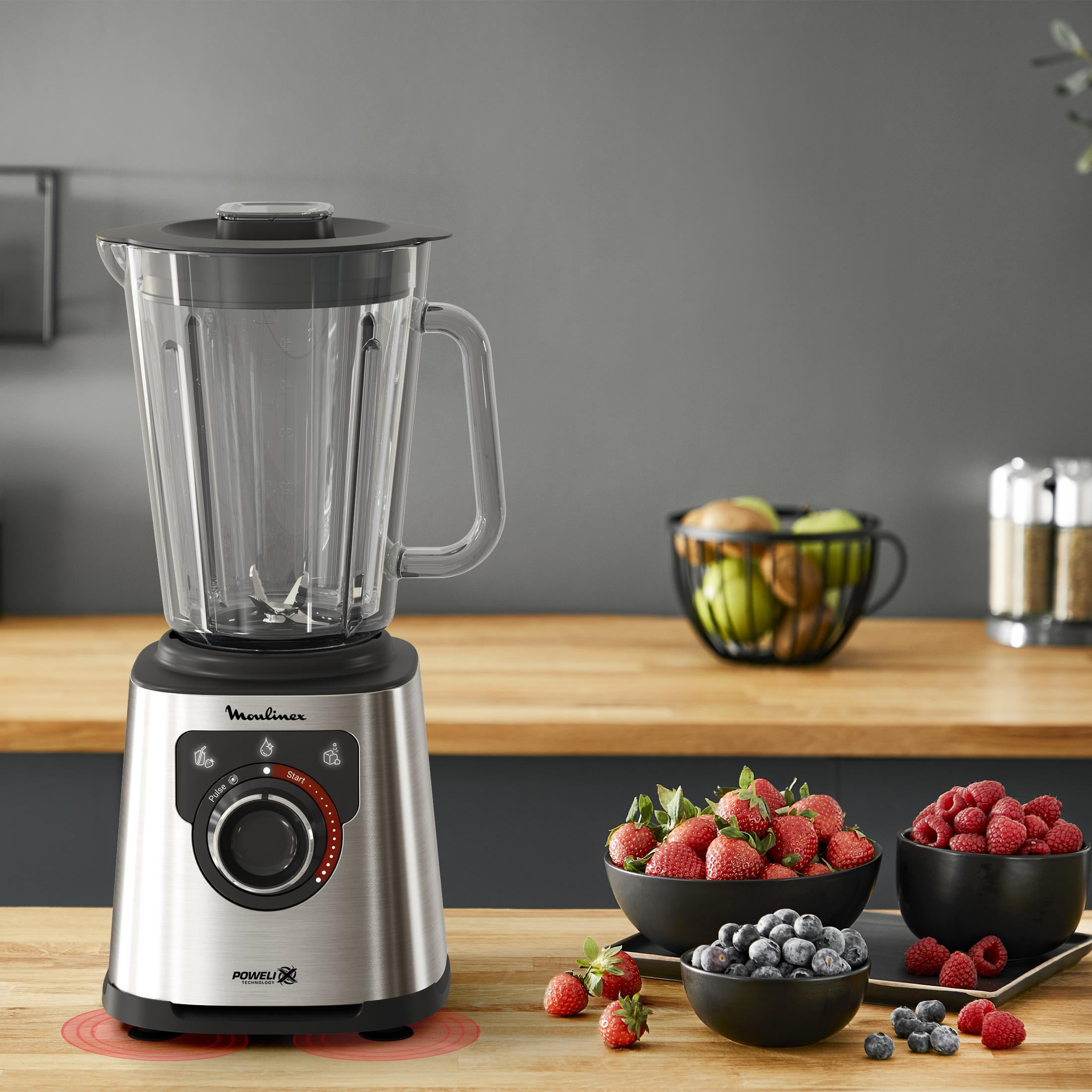 Ein silberner Mixer steht auf einem Holztisch mit Schüsseln voller Beeren.