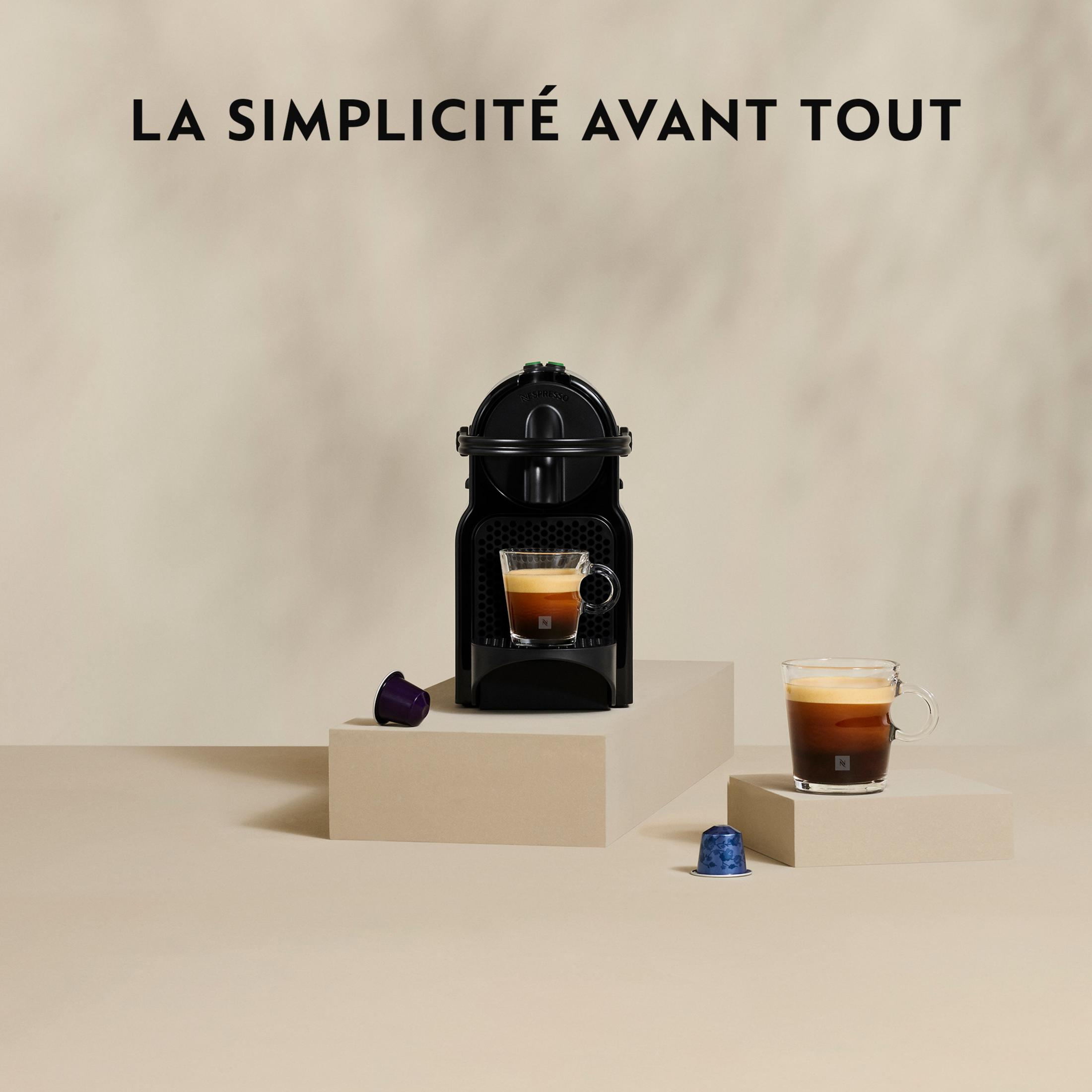 Schwarze Nespresso-Maschine mit zwei Kaffeetassen und Kapseln auf beigem Hintergrund.