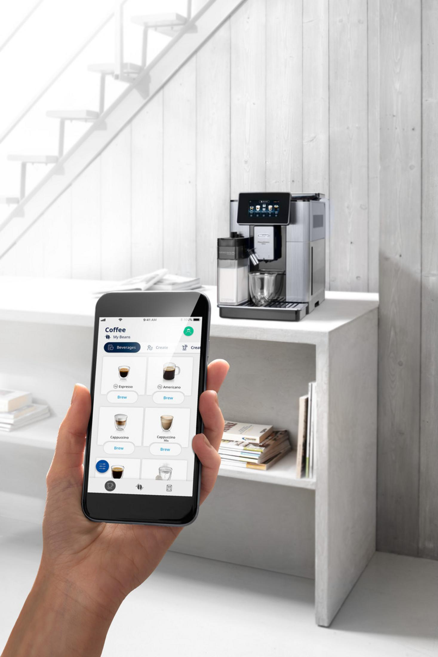 A hand holds a smartphone displaying a coffee machine app, in front of a coffee machine.