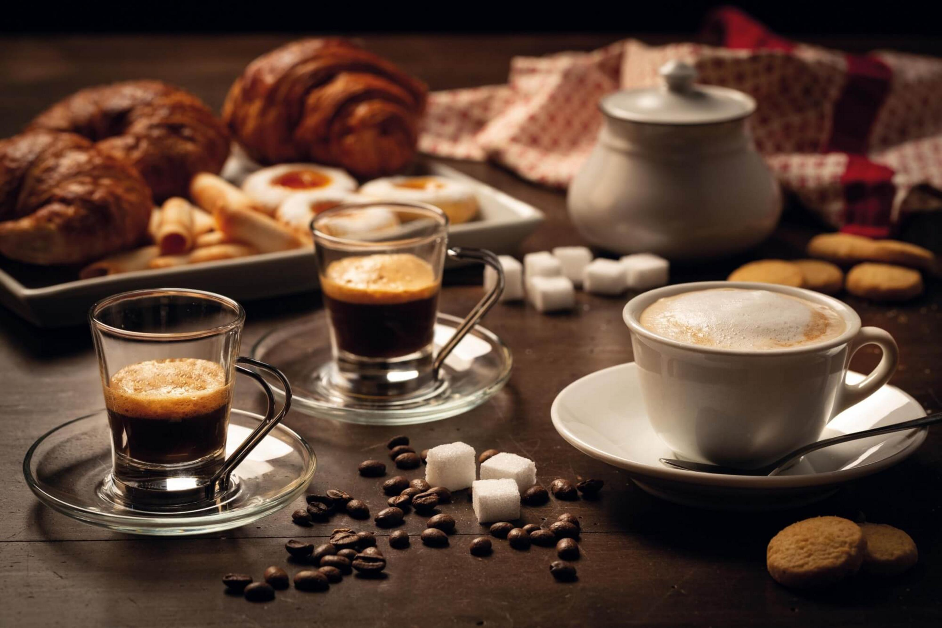 Dos espressos, un capuchino, pasteles, terrones de azúcar y granos de café en mesa madera.