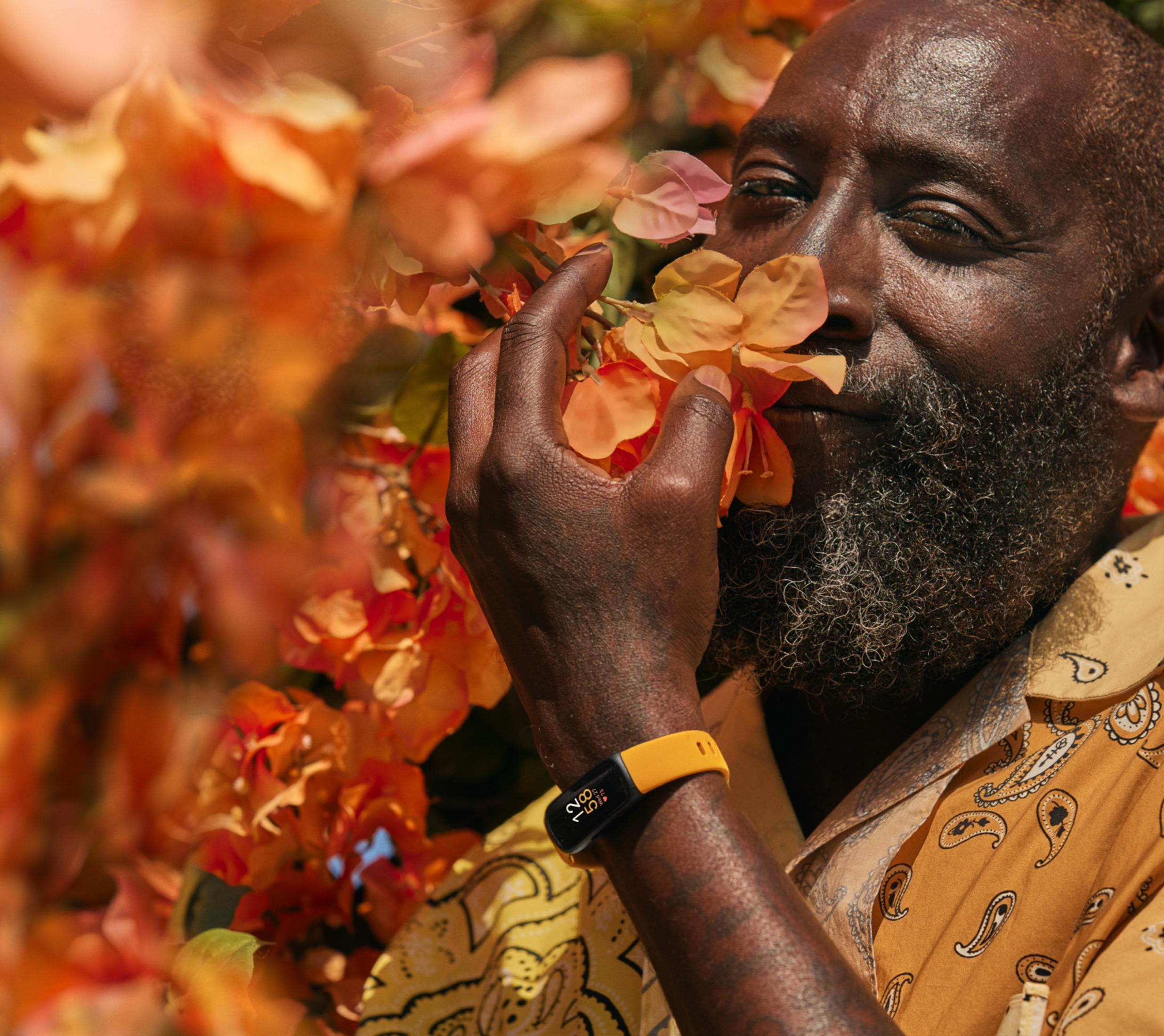 Mężczyzna z brodą wącha pomarańczowe kwiaty, nosi żółtą koszulę i zegarek fitness.