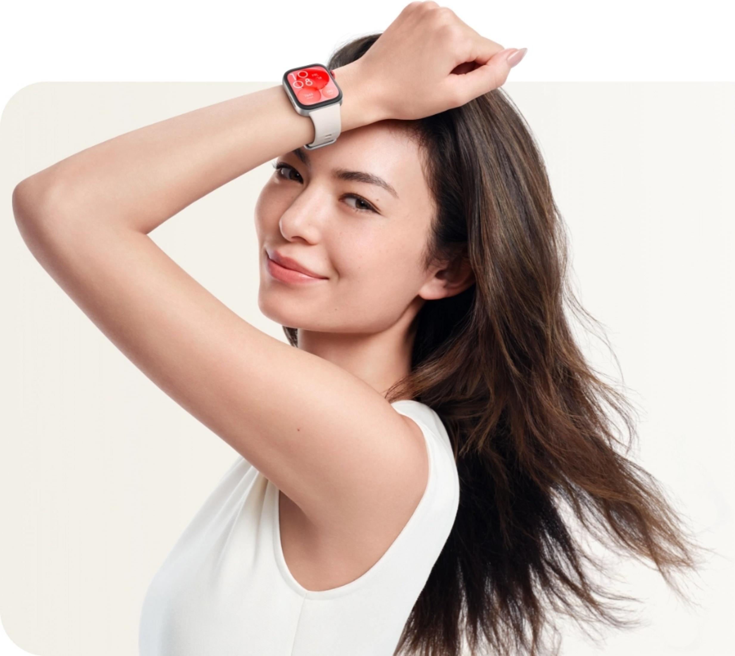 Kobieta z zegarkiem na nadgarstku, ręka nad głową, biała bluzka, jasne tło.