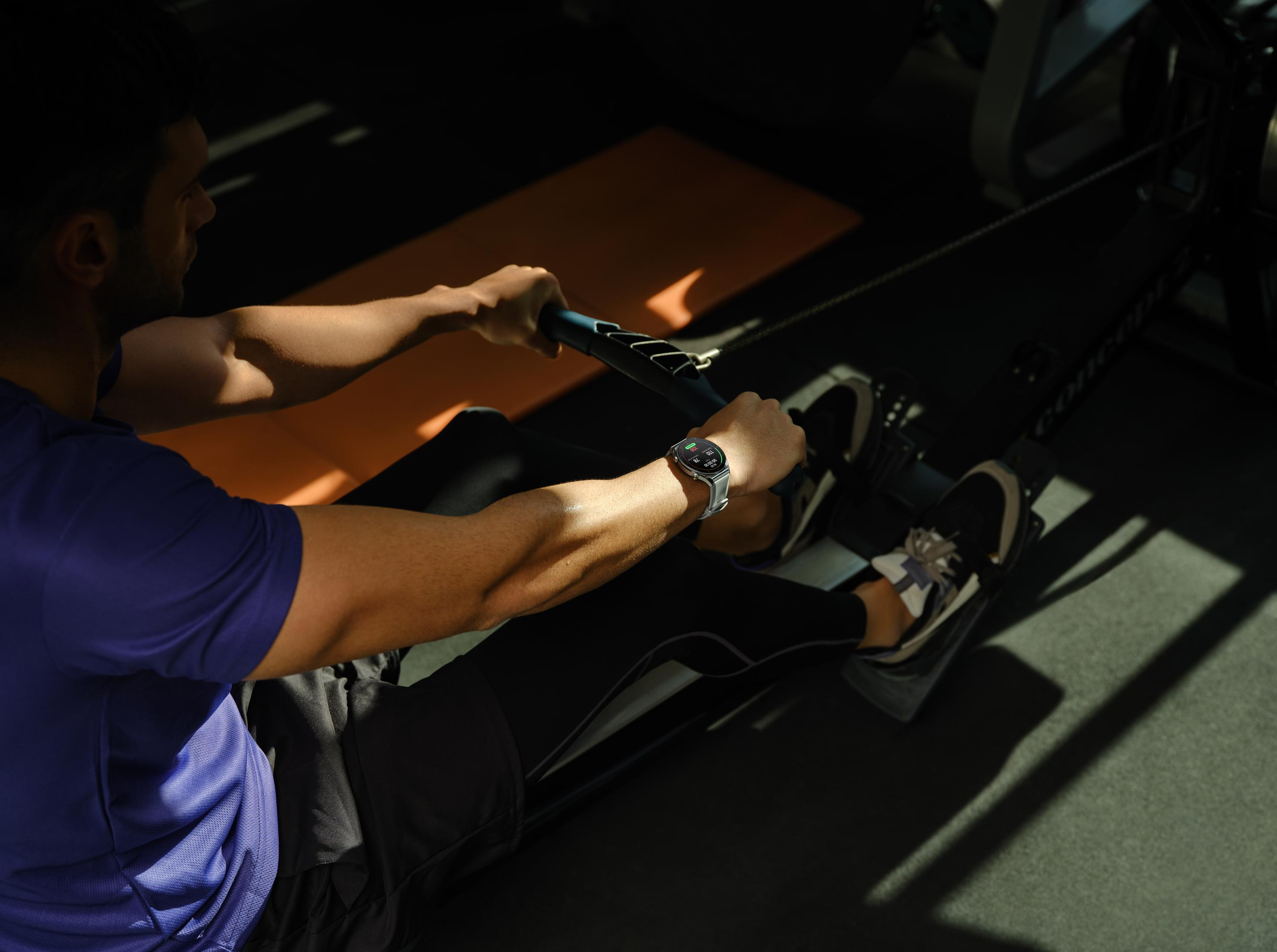 Un hombre con camiseta morada rema en una máquina en un gimnasio, con reloj.