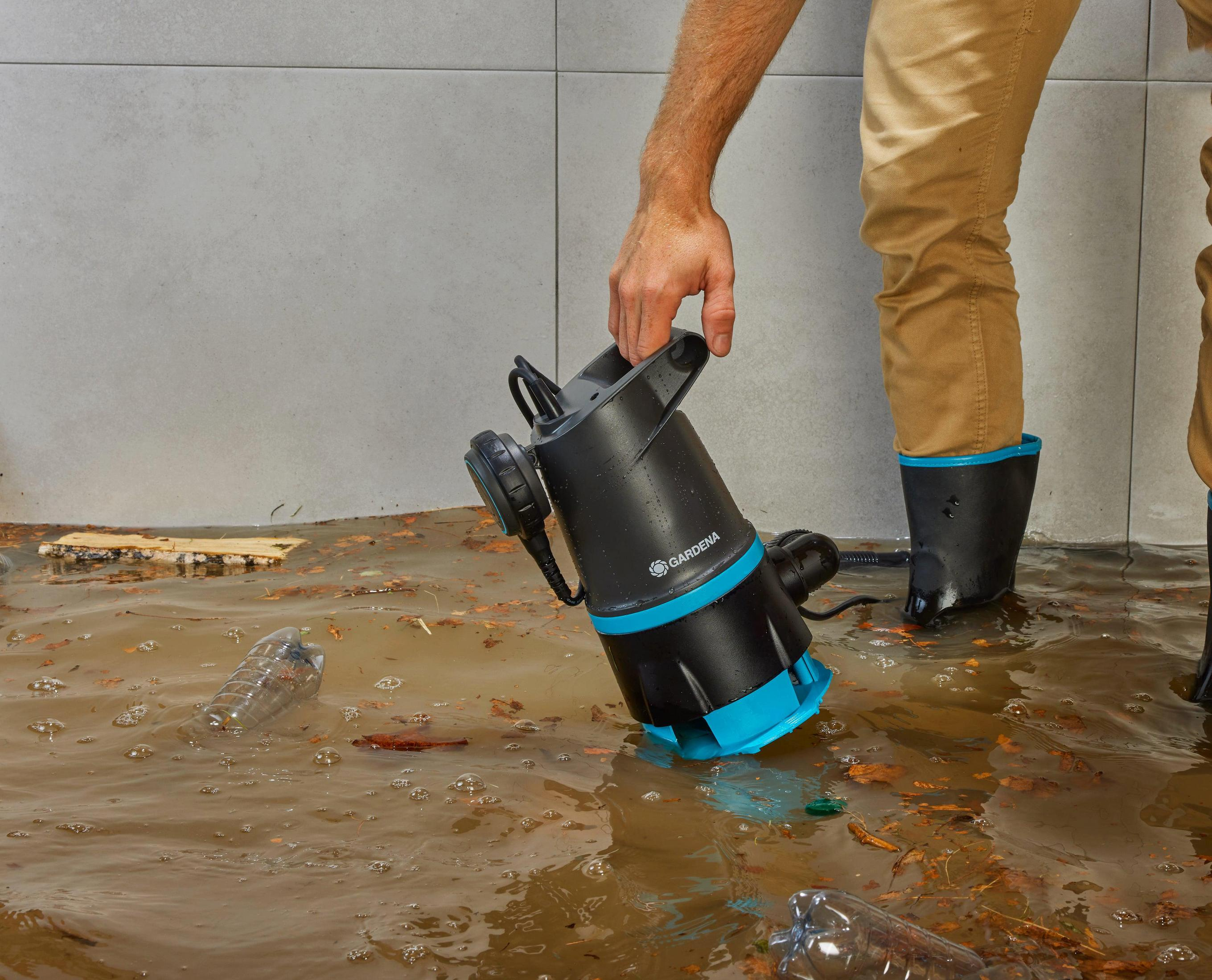 Mężczyzna w kaloszach używa pompy zanurzeniowej w błotnistej wodzie ze śmieciami.