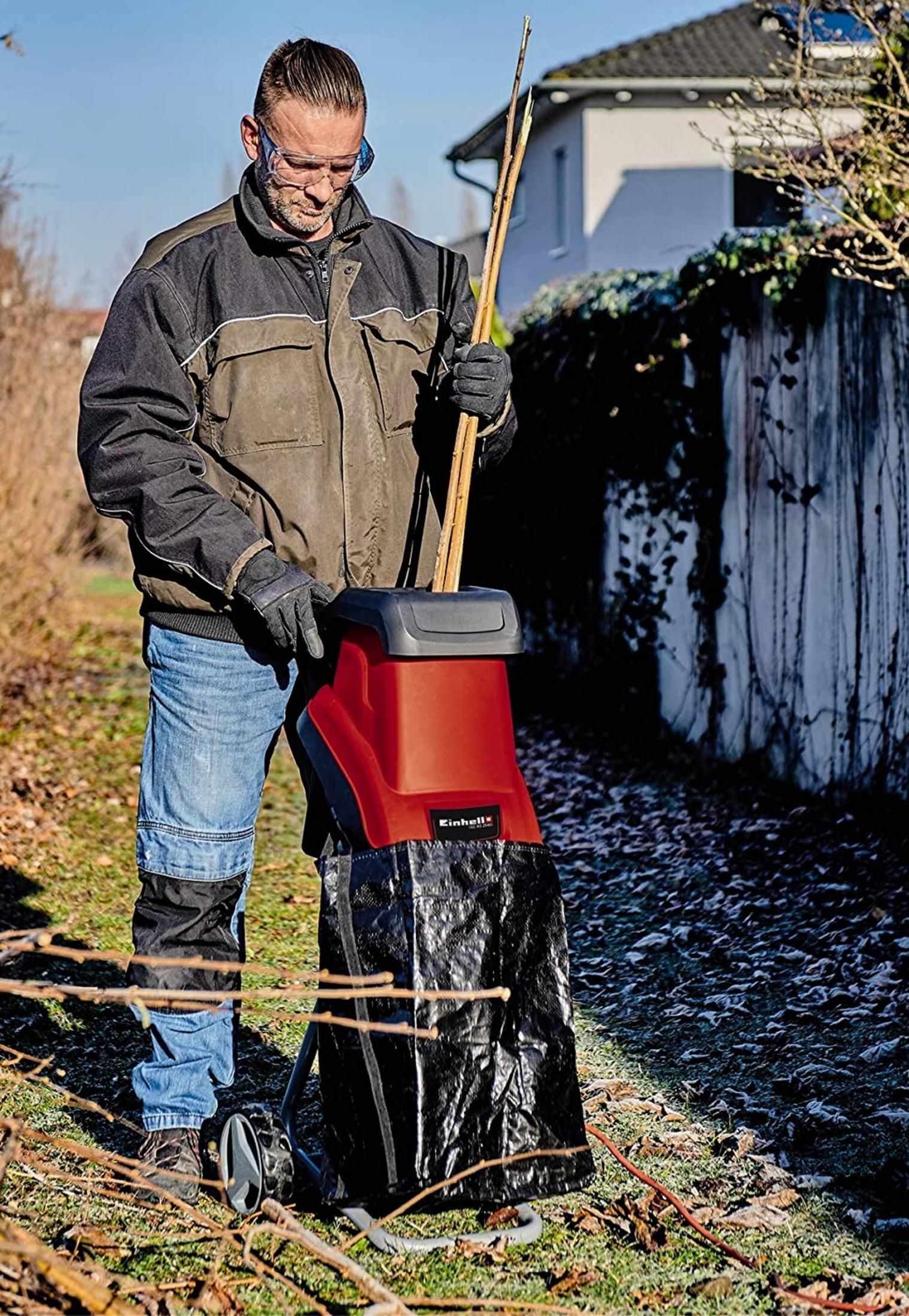 Mann mit Schutzbrille zerkleinert Zweige in einem Häcksler mit schwarzem Beutel.