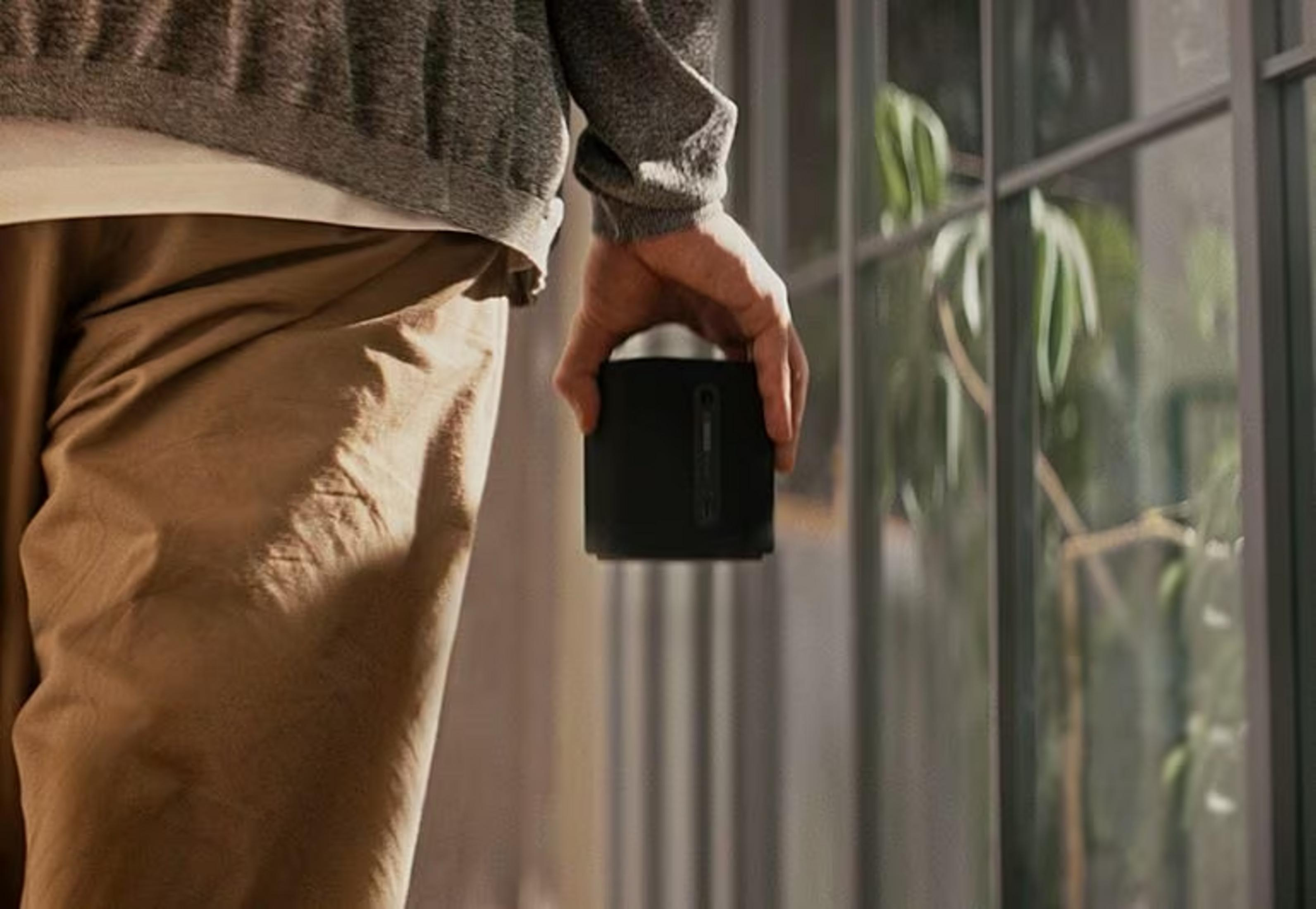 Mężczyzna trzyma czarny głośnik przed oknem z roślinami.