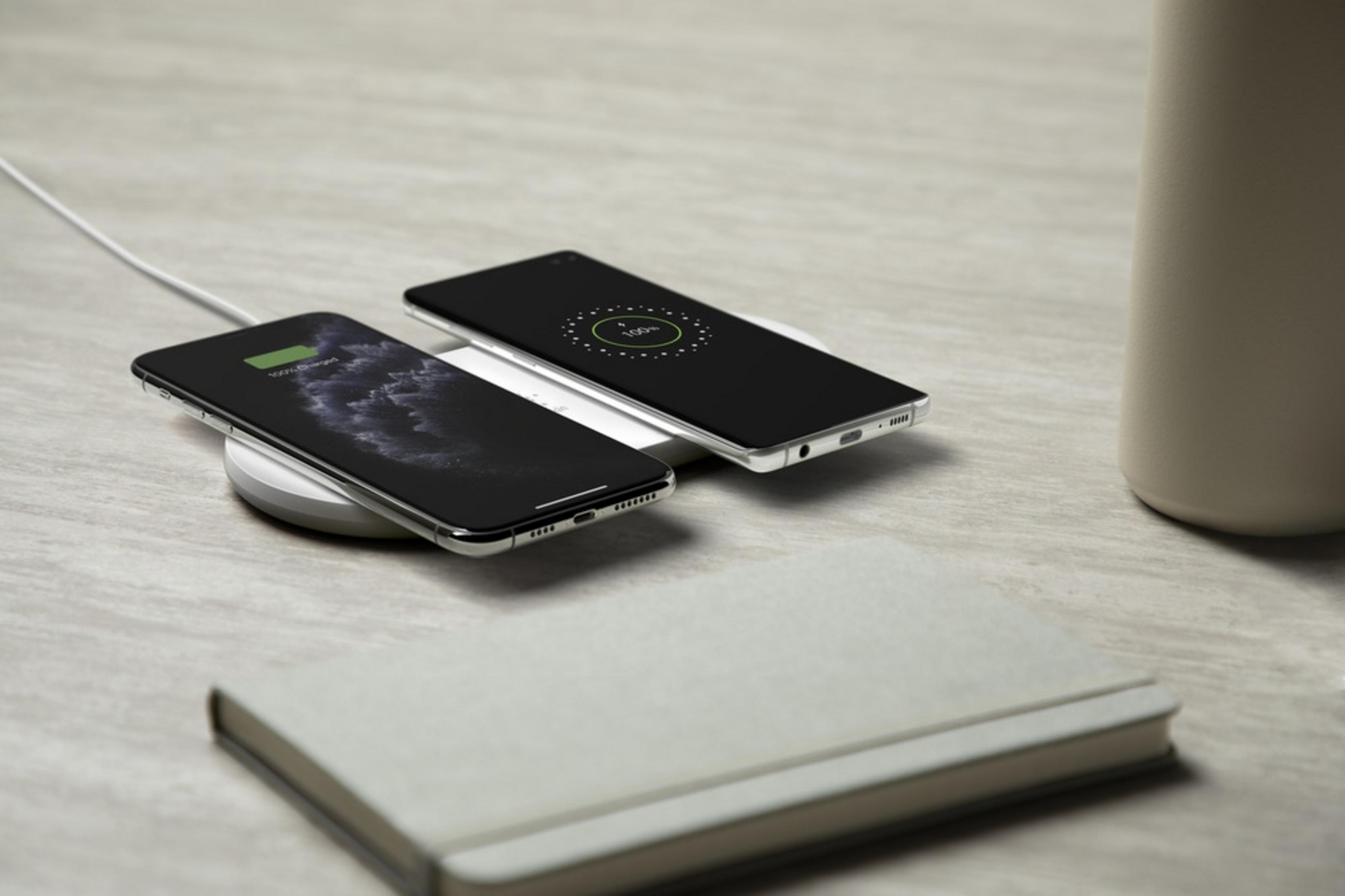 Zwei Smartphones laden auf weißen Ladestationen, daneben ein Notizbuch und eine Tasse.