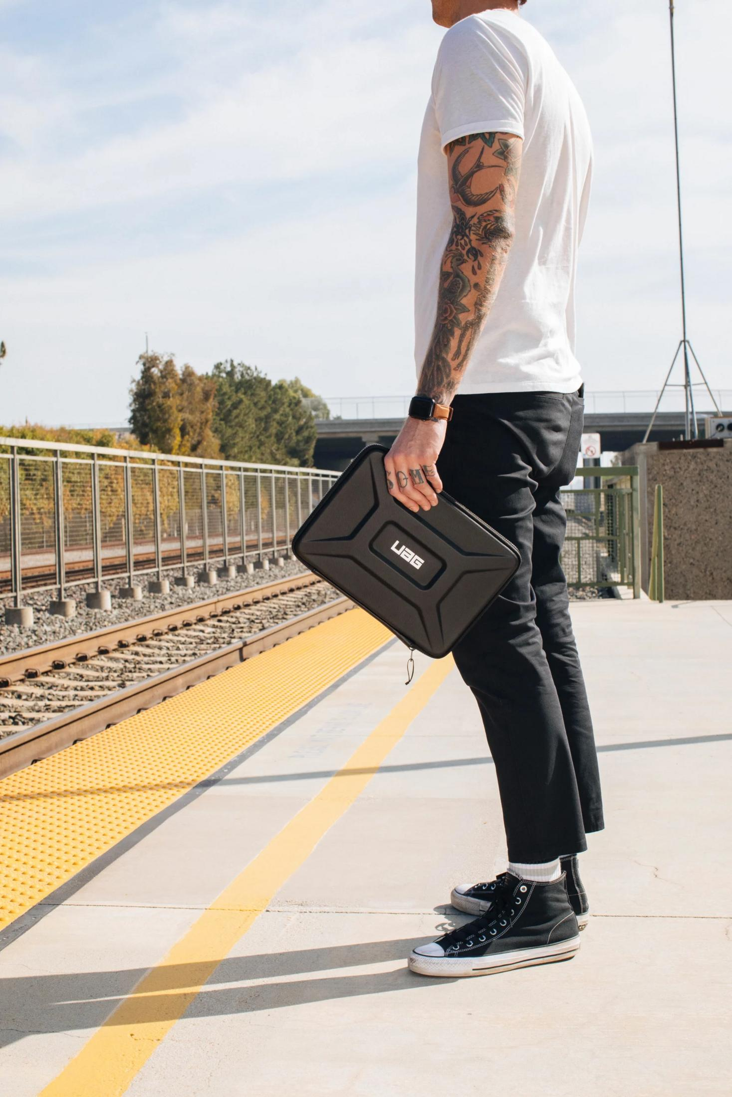 Mann mit Tattoos hält eine schwarze Tasche am Bahnsteig. Züge und Bäume im Hintergrund.