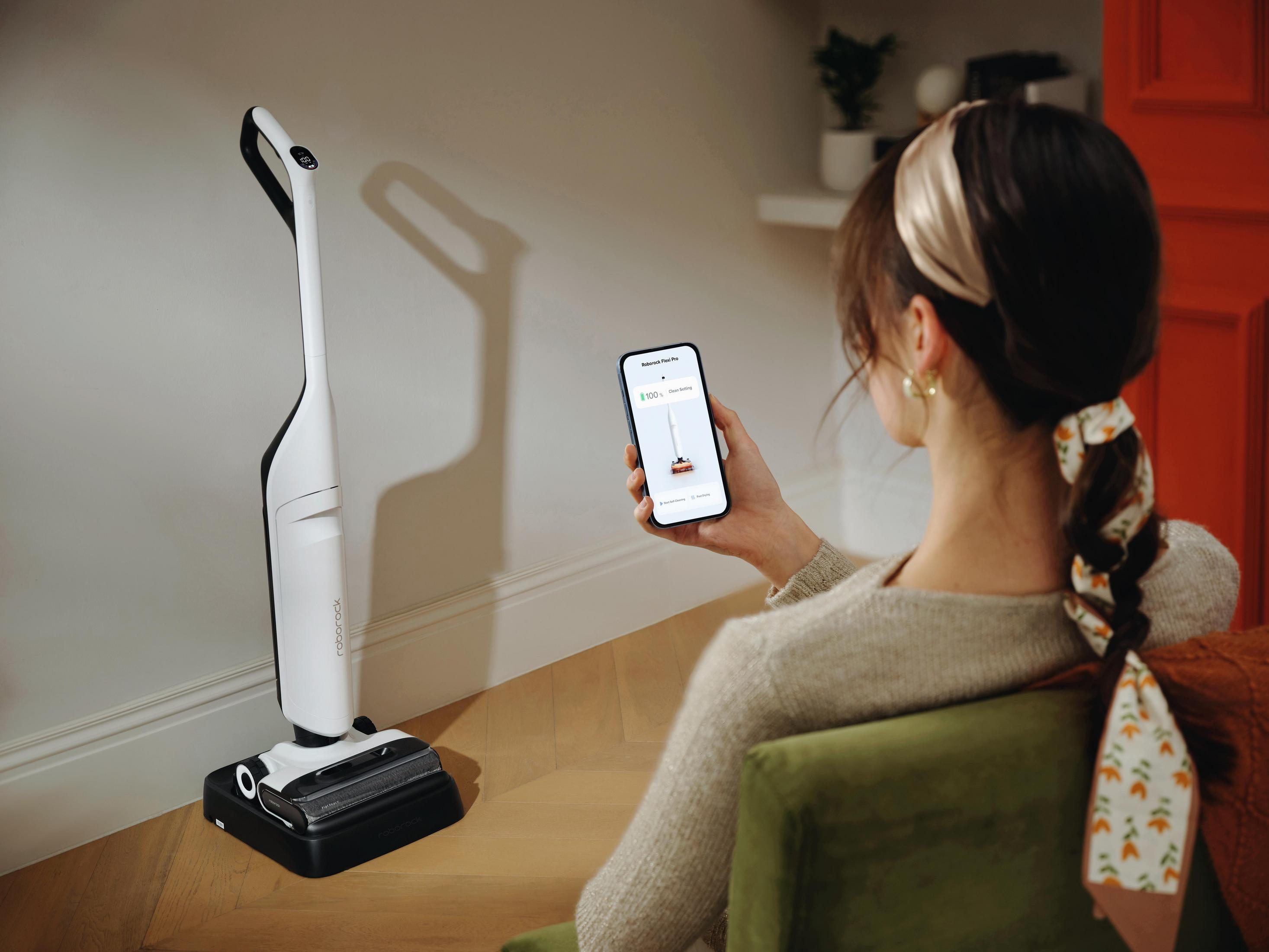Une femme tient un smartphone avec une application de nettoyage, un aspirateur est à proximité.
