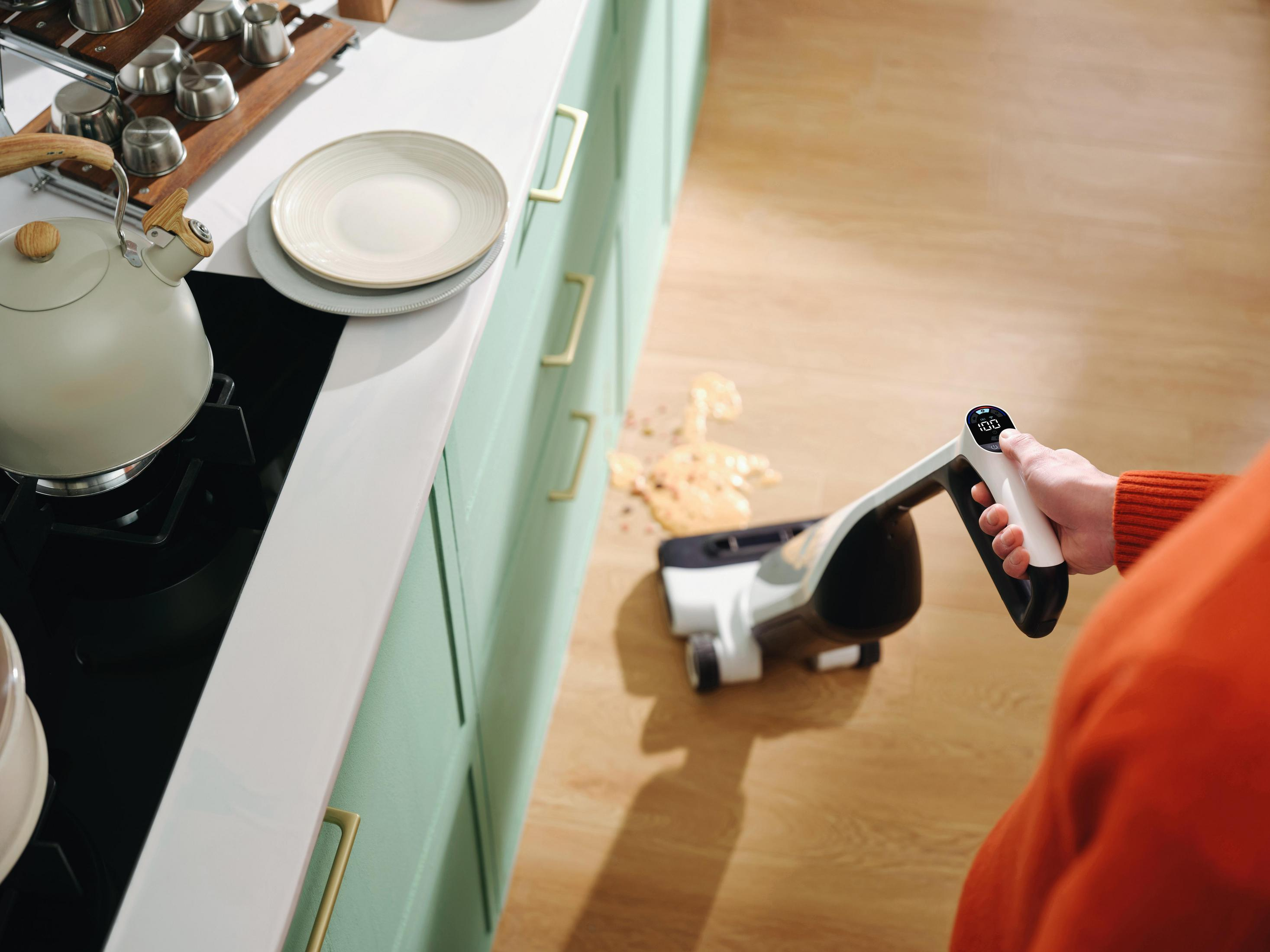 Une main utilise un aspirateur pour aspirer de la nourriture renversée sur un plancher en bois.