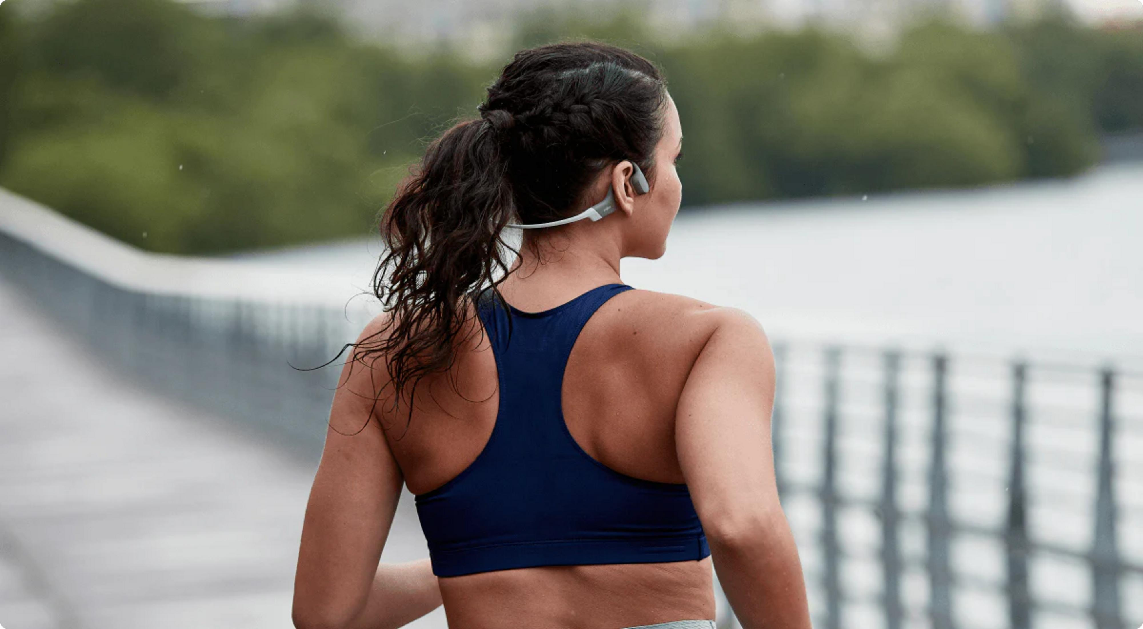 Biegaczka ze słuchawkami na moście, widziana od tyłu, z zieloną roślinnością.