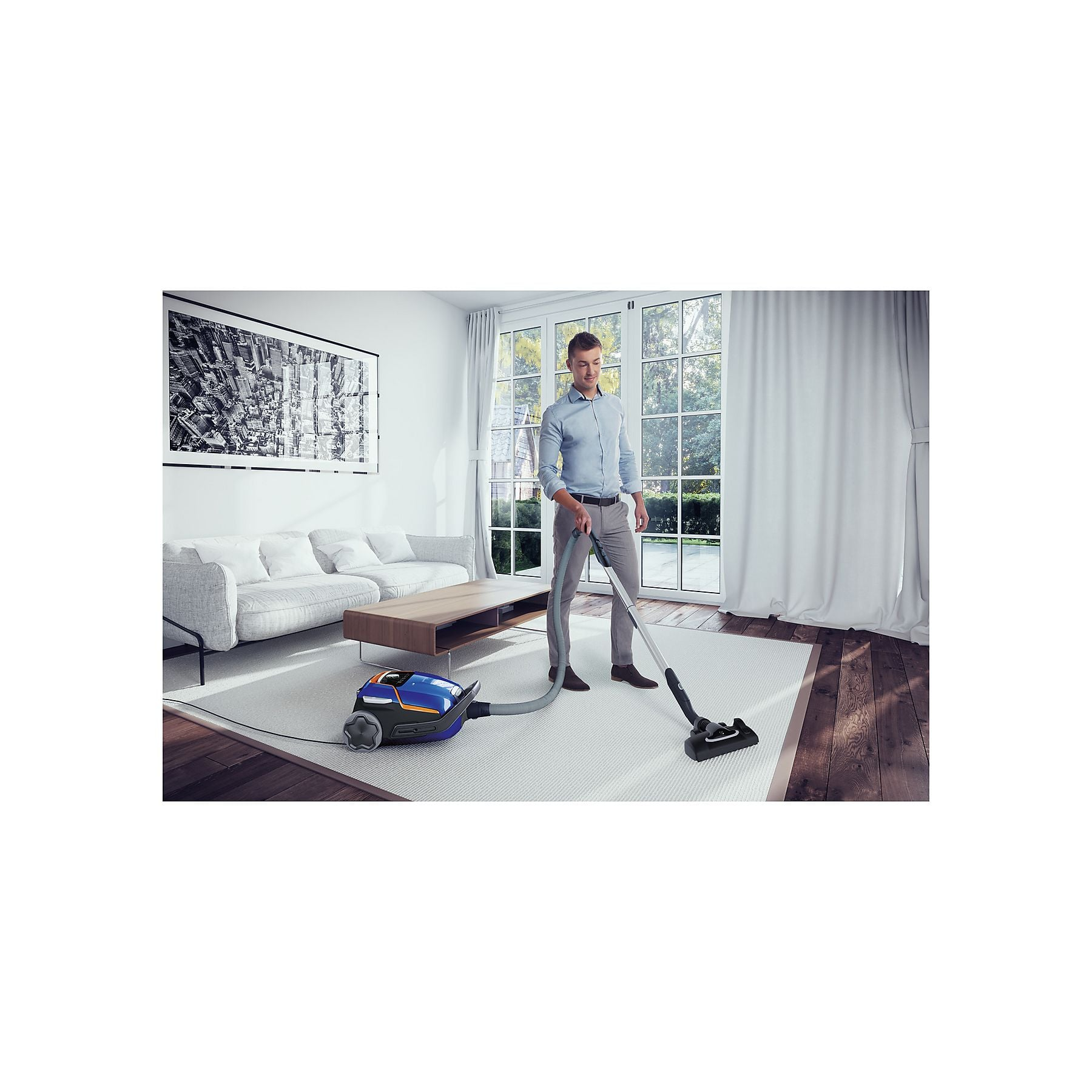 Mężczyzna odkurza dywan niebieskim odkurzaczem obok sofy i stolika.