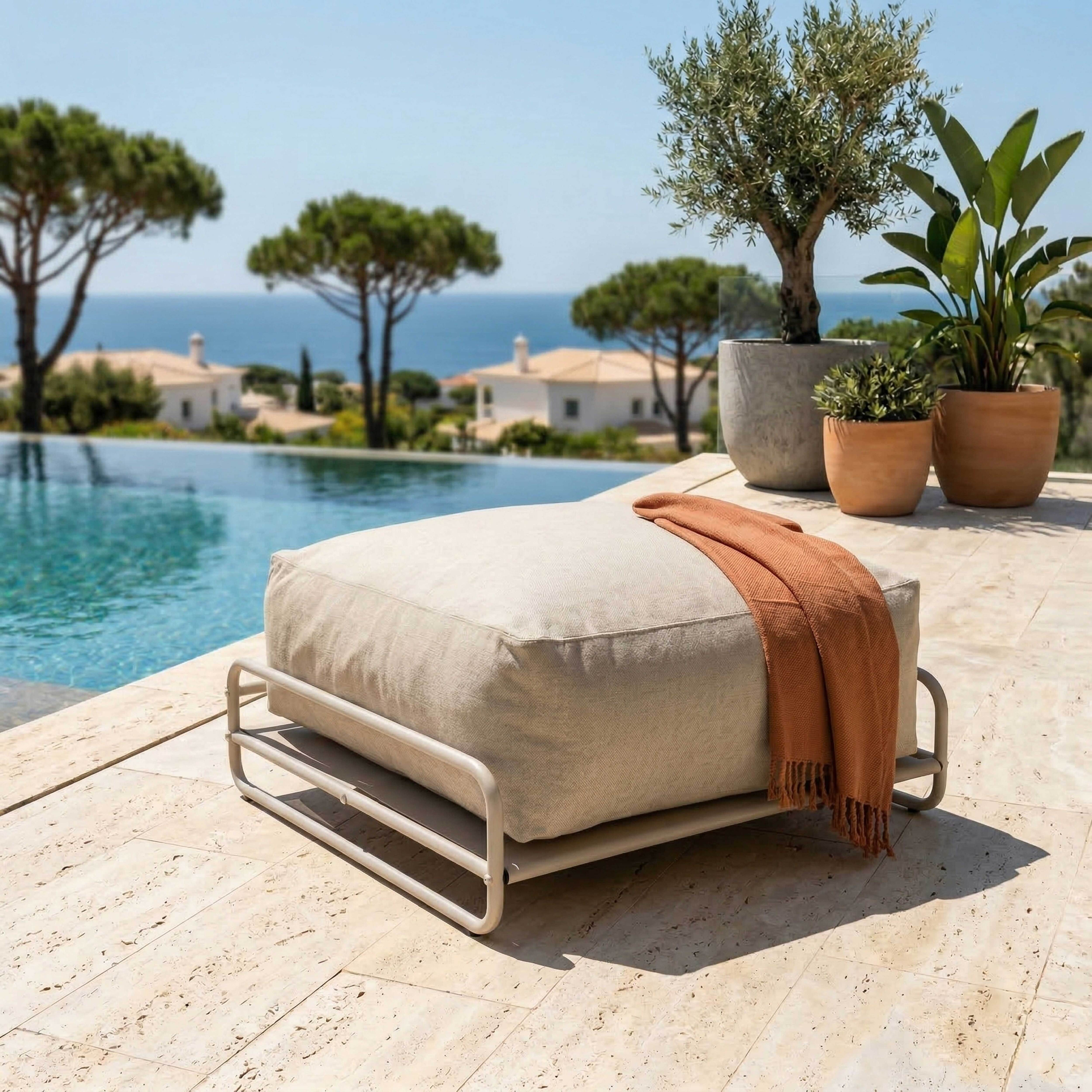 Ein beiger Hocker mit orangefarbener Decke neben einem Pool mit Blick auf das Meer.