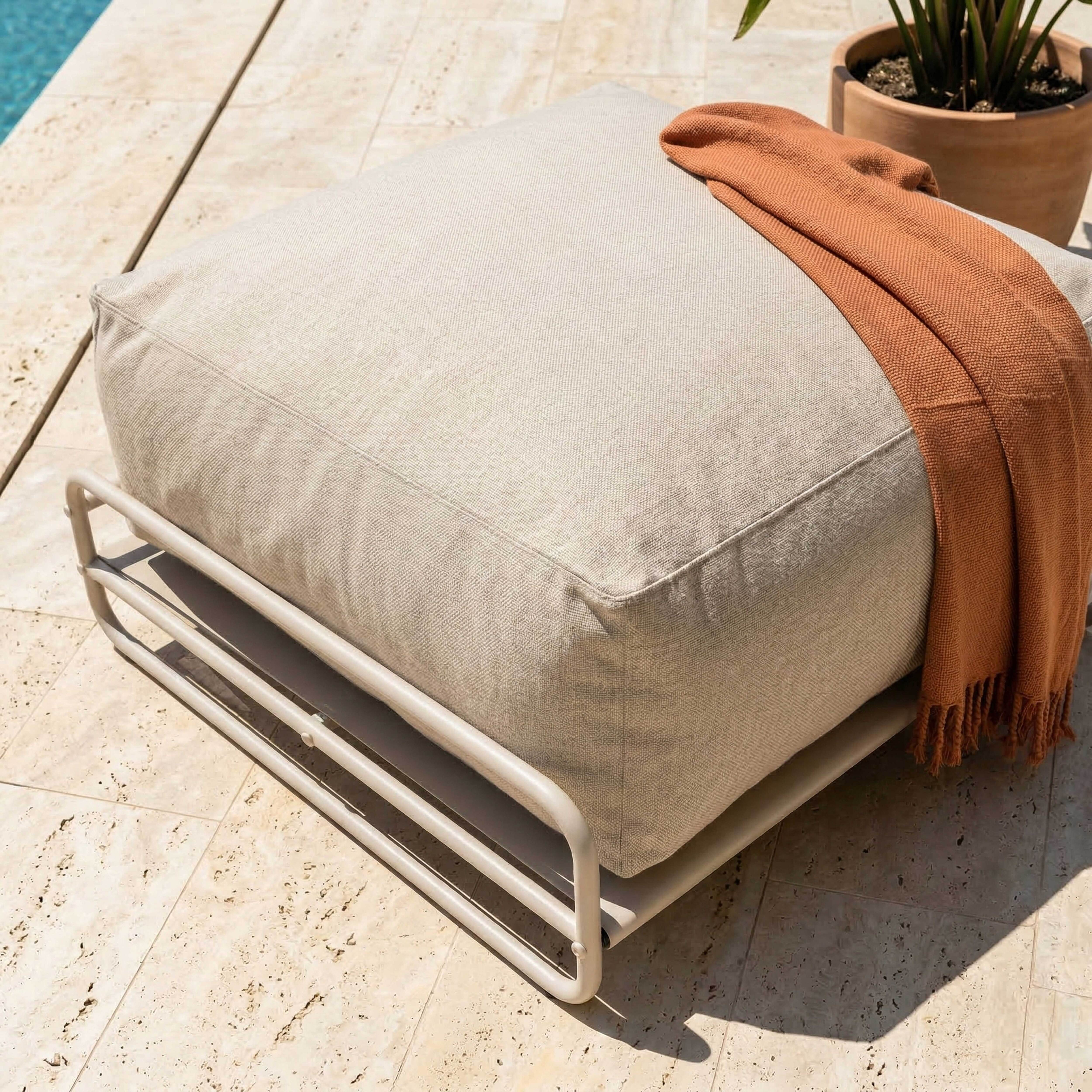Ein beiger Hocker mit orangefarbener Decke auf Steinboden neben einem Pool.