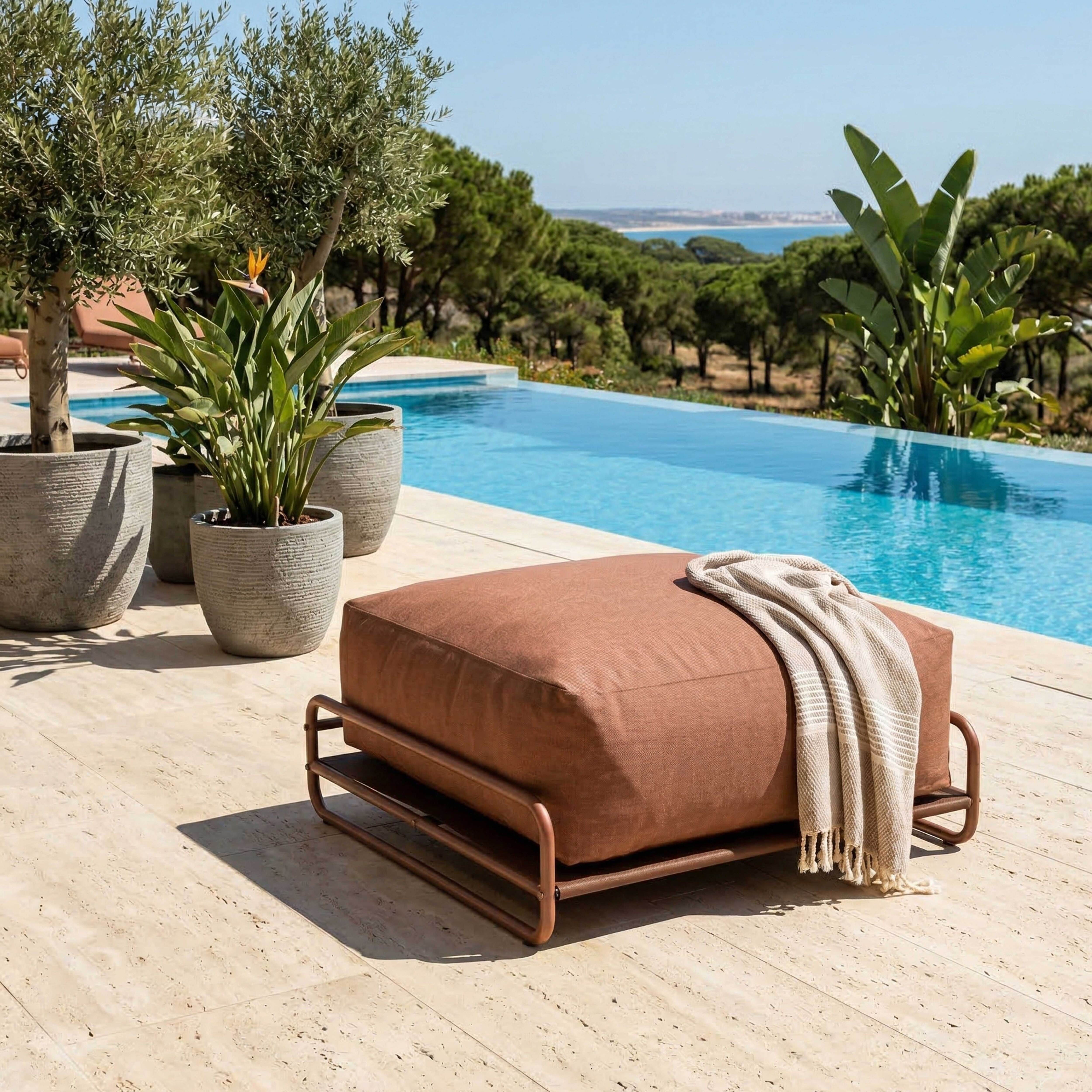 Ein brauner Hocker mit Decke neben einem Pool, mit Bäumen und dem Meer im Hintergrund.
