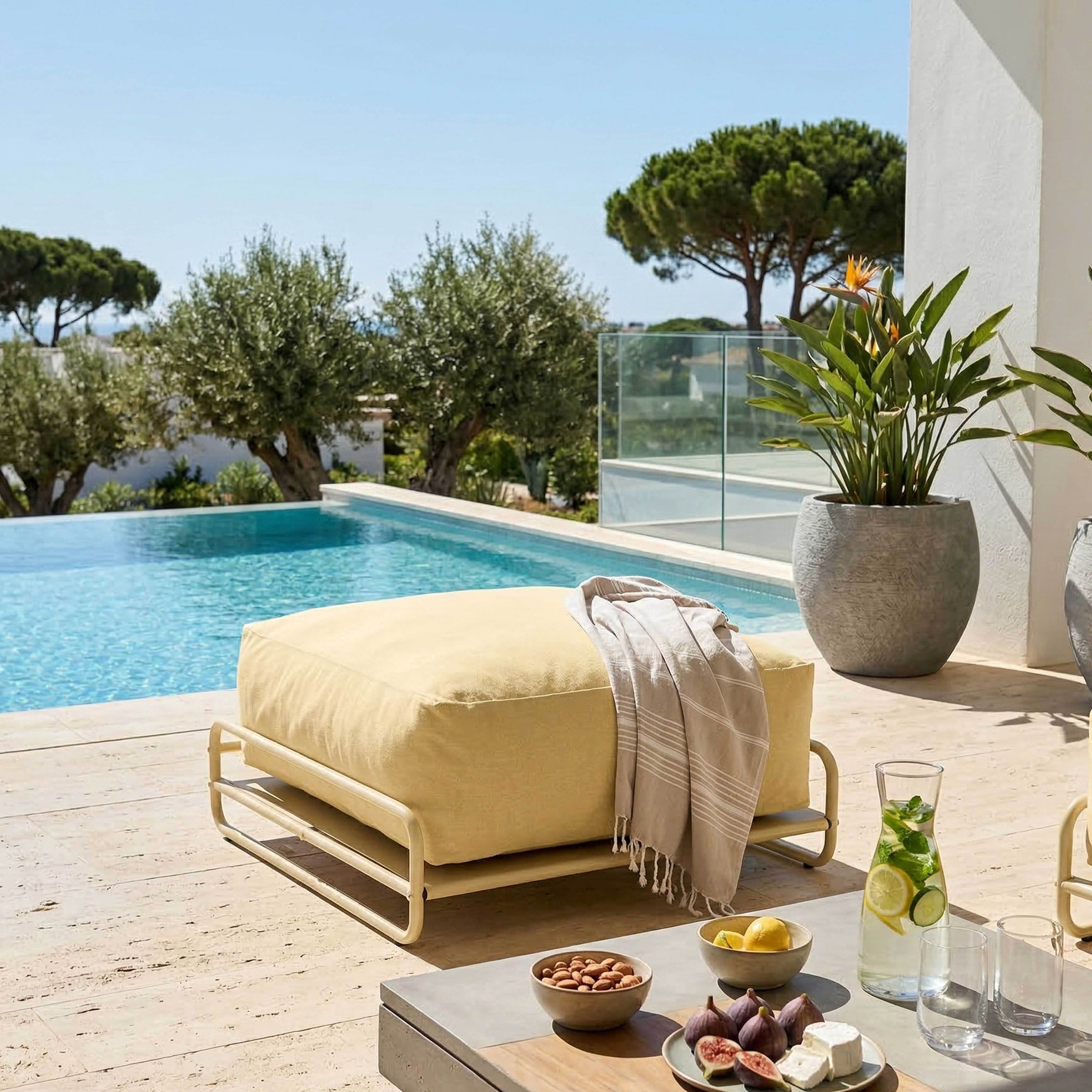 Ein gelber Hocker am Pool mit Olivenbäumen und einem Haus. Erfrischungen auf dem Tisch.