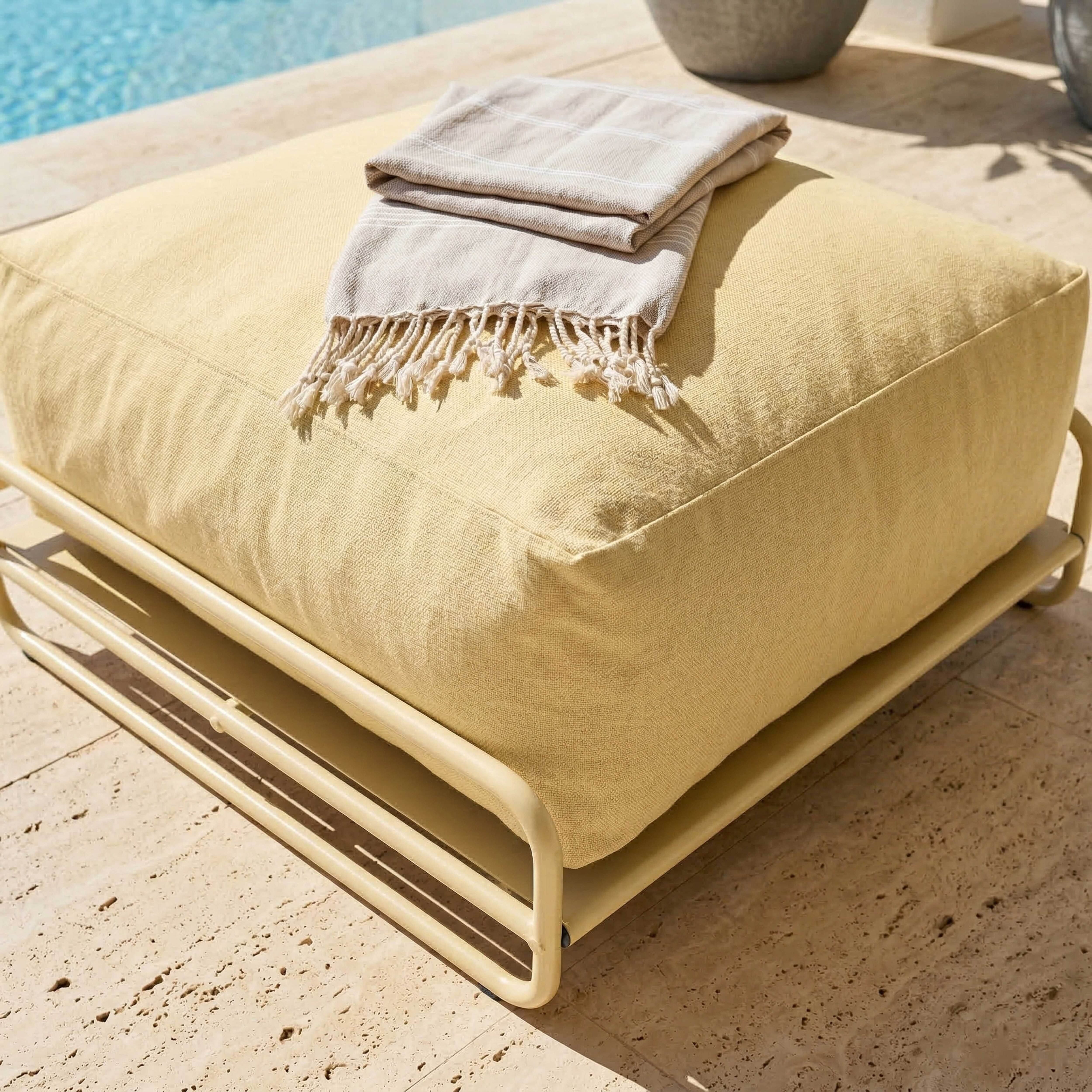 Ein gelbes Bodenkissen mit beige'm Handtuch daneben am Pool.