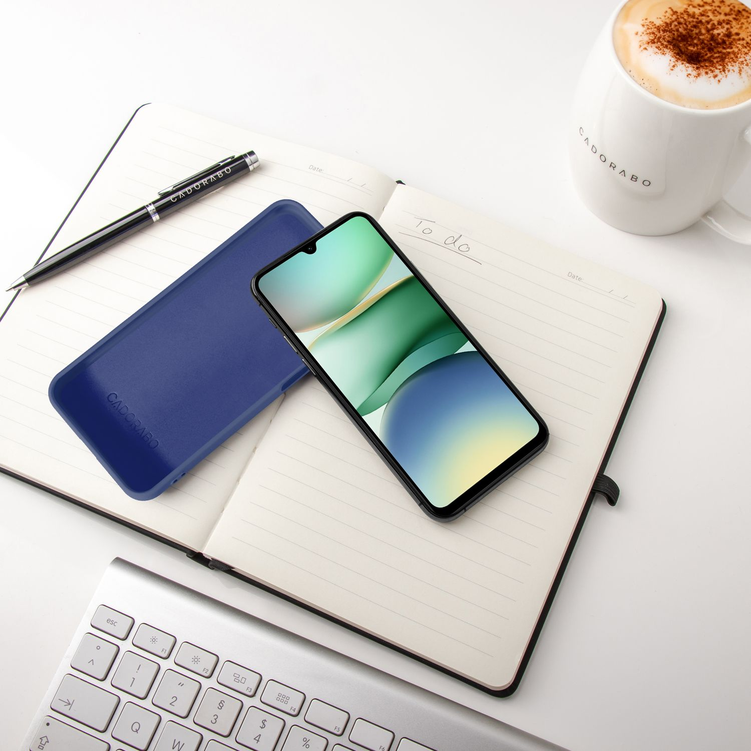 Ein Smartphone liegt auf einem Notizbuch neben einer blauen Hülle, Stift, Tasse Kaffee und Tastatur.