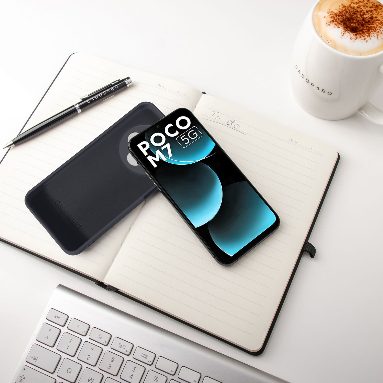Ein Smartphone, Notizbuch, Stift, Tastatur und Kaffeetasse auf einem weißen Schreibtisch.