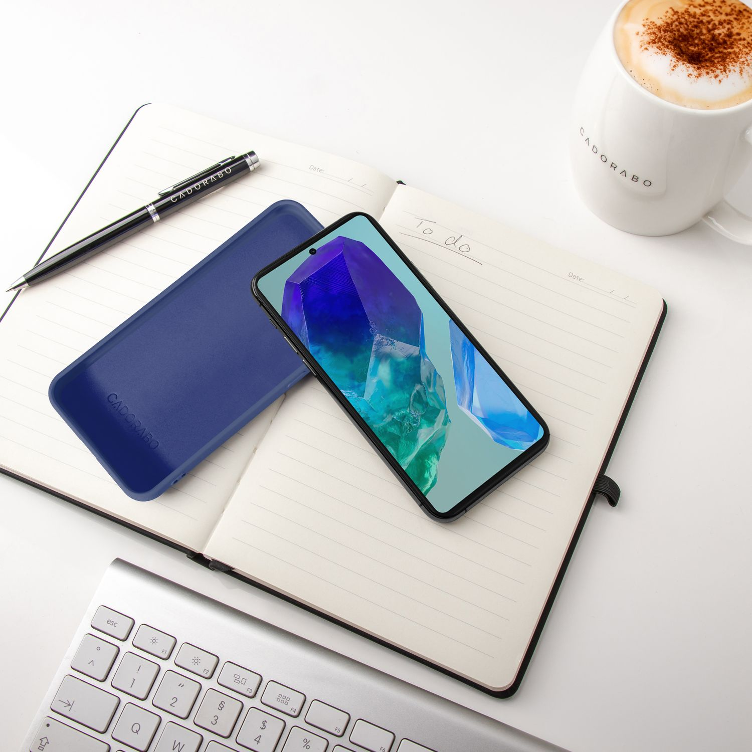 Ein Smartphone liegt auf einem Notizbuch neben einer blauen Hülle und einer Tasse Kaffee.