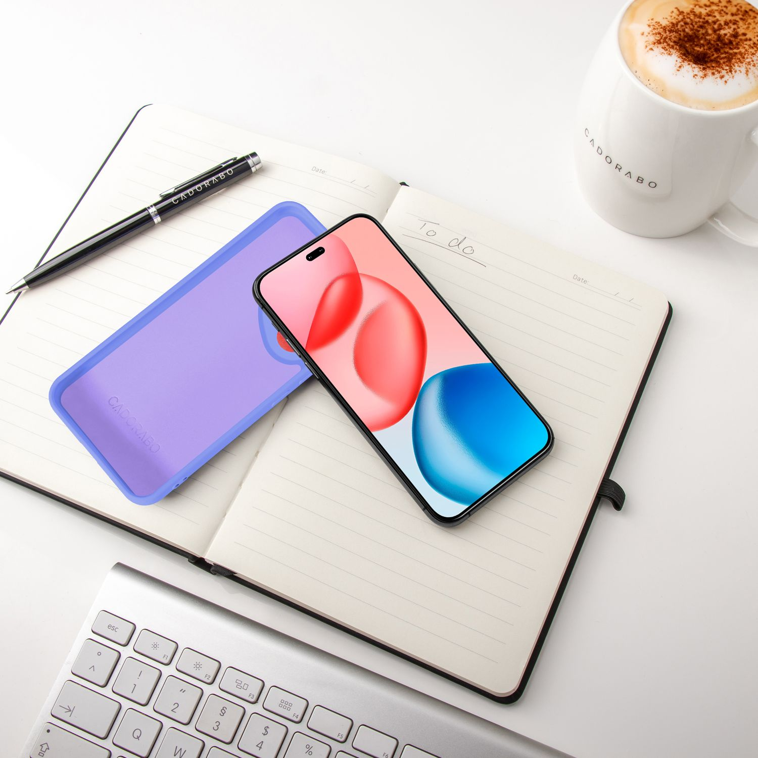 Ein Notizbuch mit Telefon, lila Hülle, Stift, Tastatur und Kaffeetasse auf einem weißen Schreibtisch.