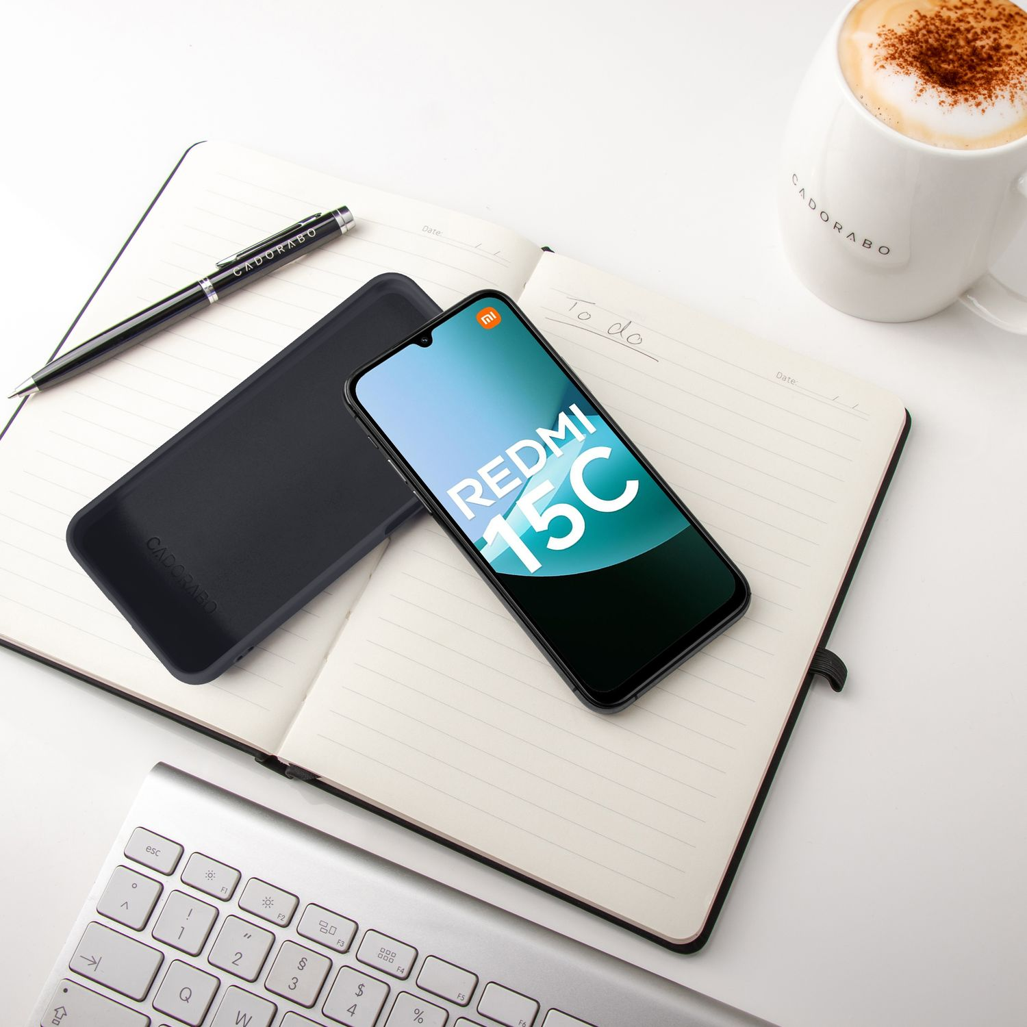 Ein Smartphone, Notizbuch, Stift, Tastatur und eine Kaffeetasse auf einem weißen Tisch.