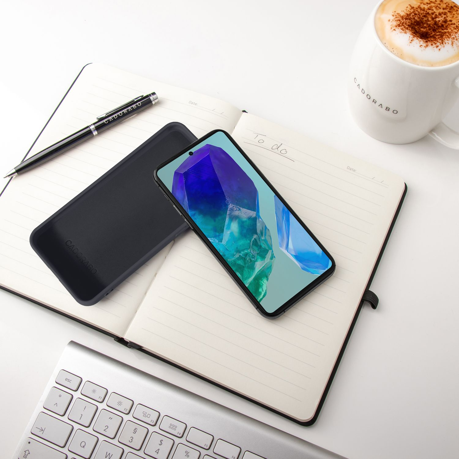 Ein Smartphone, ein Notizbuch, ein Stift, eine Tastatur und eine Kaffeetasse auf einem weißen Tisch.
