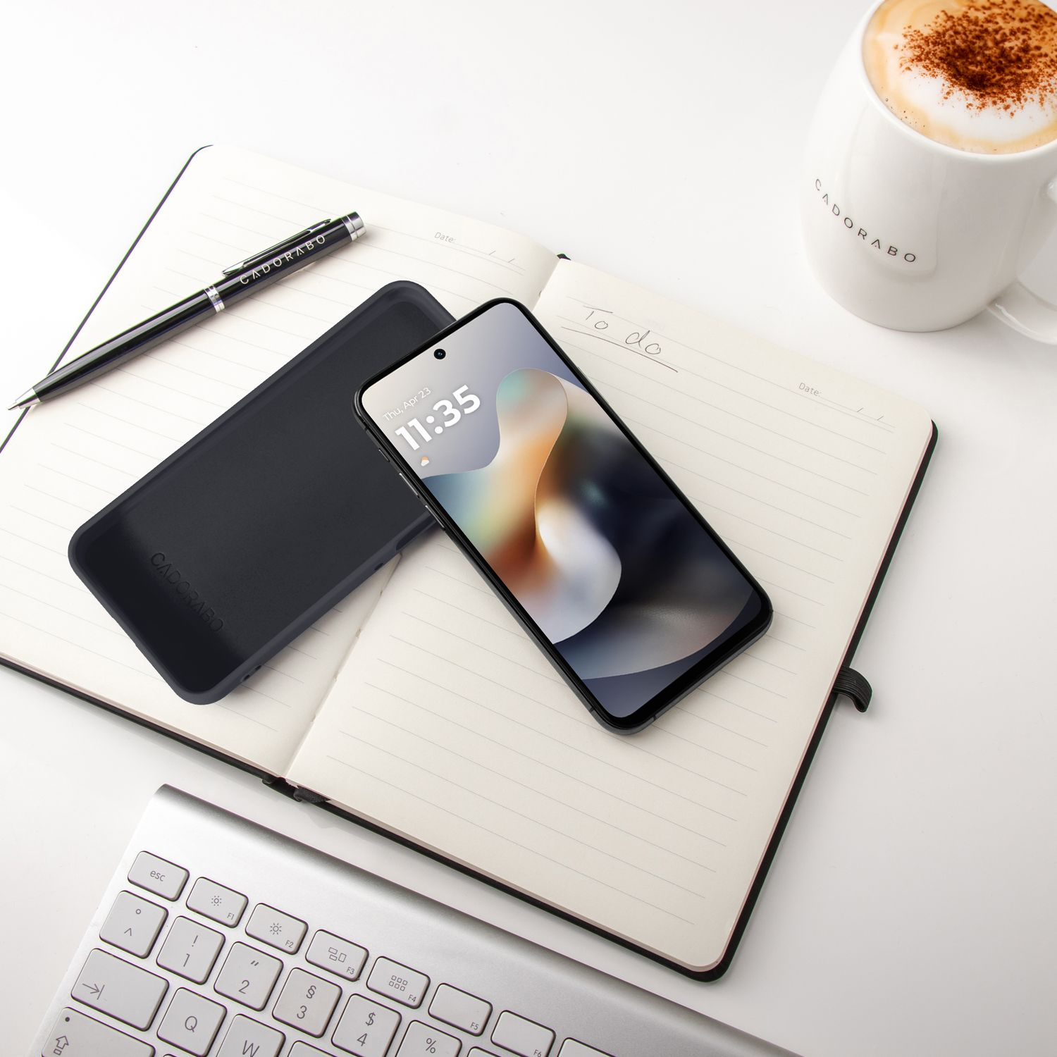 Ein schwarzes Handy liegt neben einem Stift und einer Kaffeetasse auf einem Notizbuch.