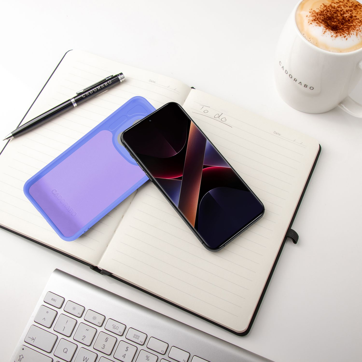 Ein Smartphone, ein Notizbuch, ein Stift, eine Tastatur und eine Kaffeetasse liegen auf einem weißen Tisch.