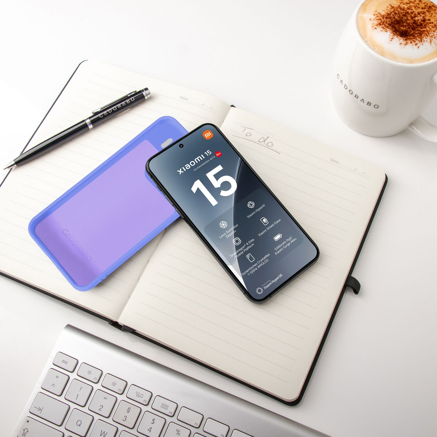 Ein Handy, eine lila Hülle, ein Notizbuch, ein Stift und eine Kaffeetasse liegen auf einem Schreibtisch.