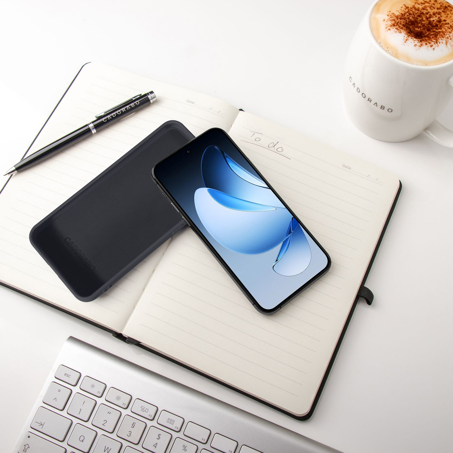 Ein Handy, Notizbuch, Stift, Tastatur und Kaffee auf einem Schreibtisch.
