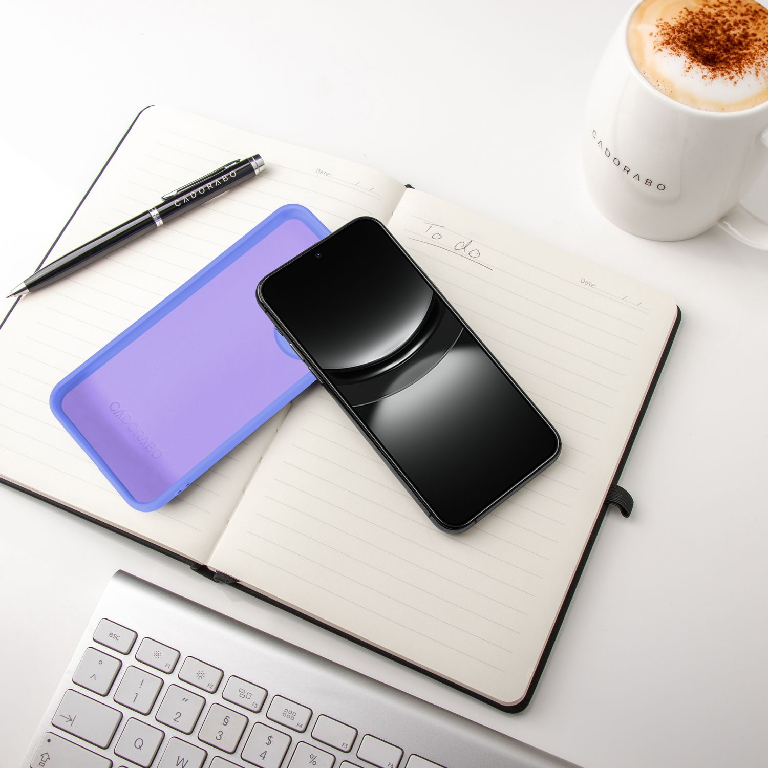 Schwarzes Smartphone, lila Hülle, Notizbuch, Stift und Kaffee auf weißem Hintergrund.