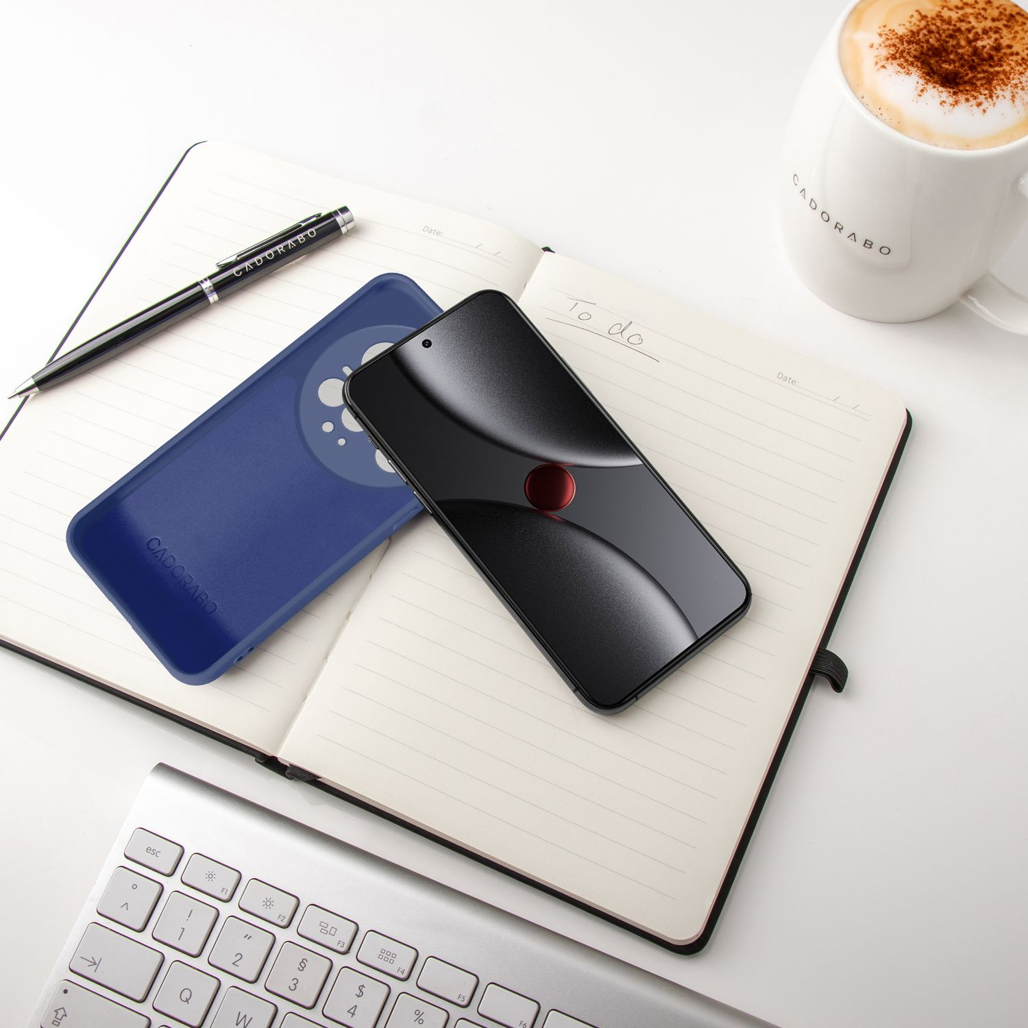 Ein schwarzes Smartphone liegt auf einem Notizbuch neben einer blauen Hülle, Stift und Kaffee.