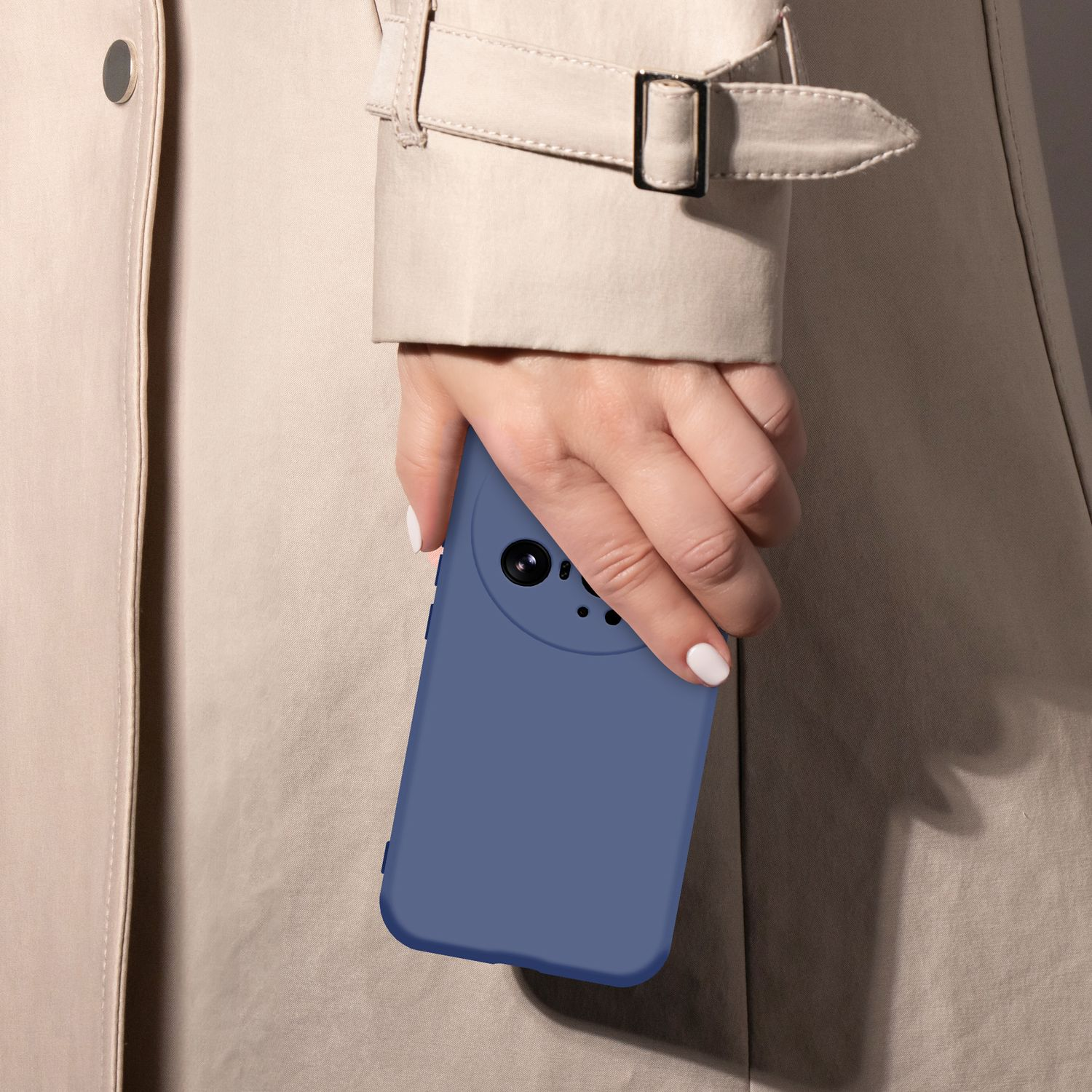 Eine Hand mit blauen Nägeln hält ein blaues Handy vor einem beigefarbenen Trenchcoat.