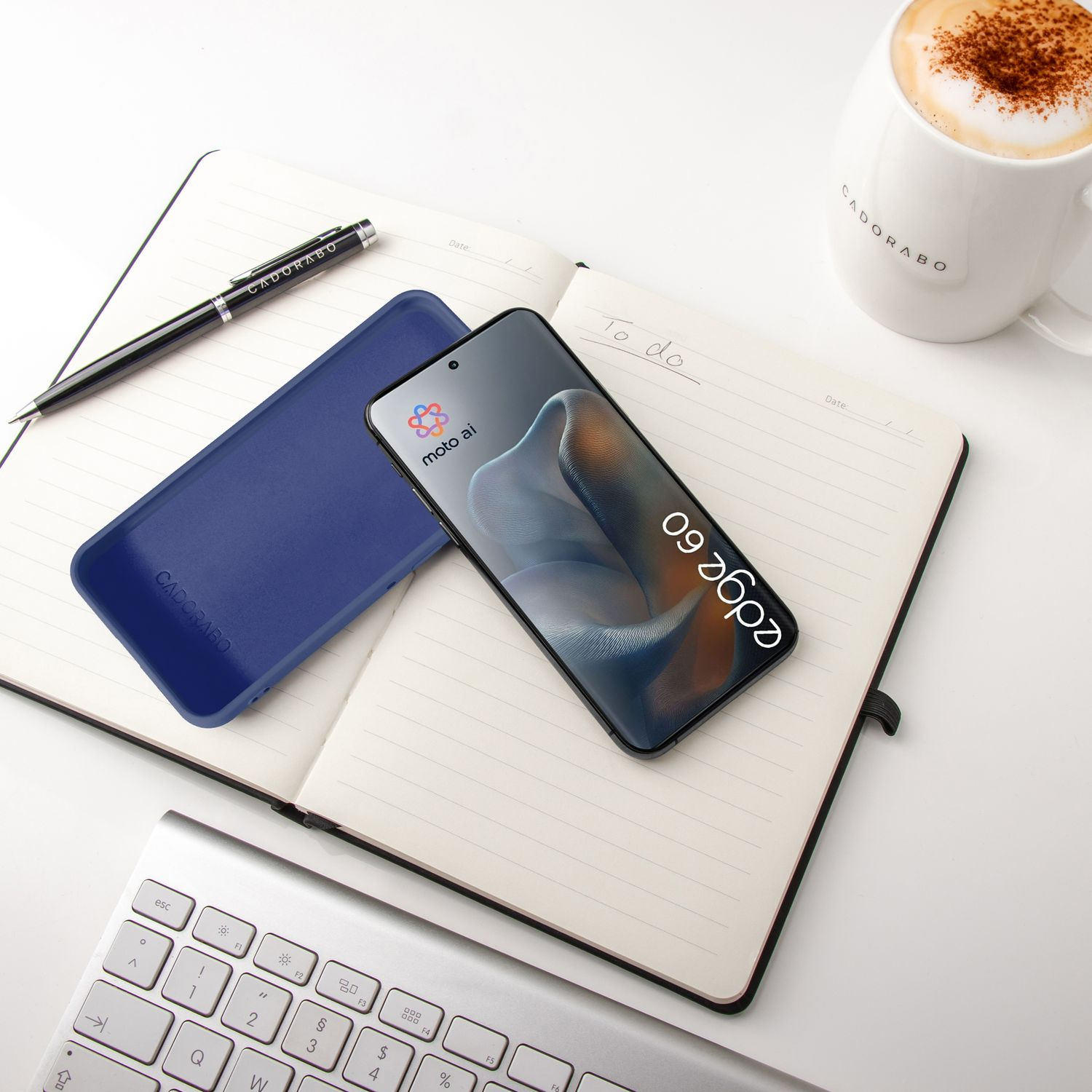 Ein Smartphone, Notizbuch, Stift, Tastatur und eine Kaffeetasse auf einem weißen Tisch.