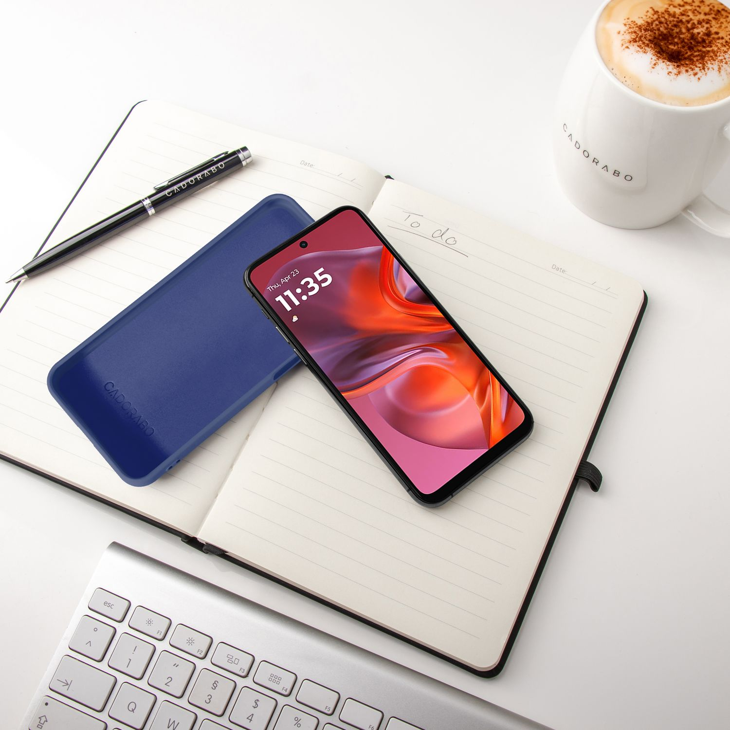 Ein aufgeschlagenes Notizbuch mit einem blauen Handycover, Stift, Handy und Kaffeebecher.
