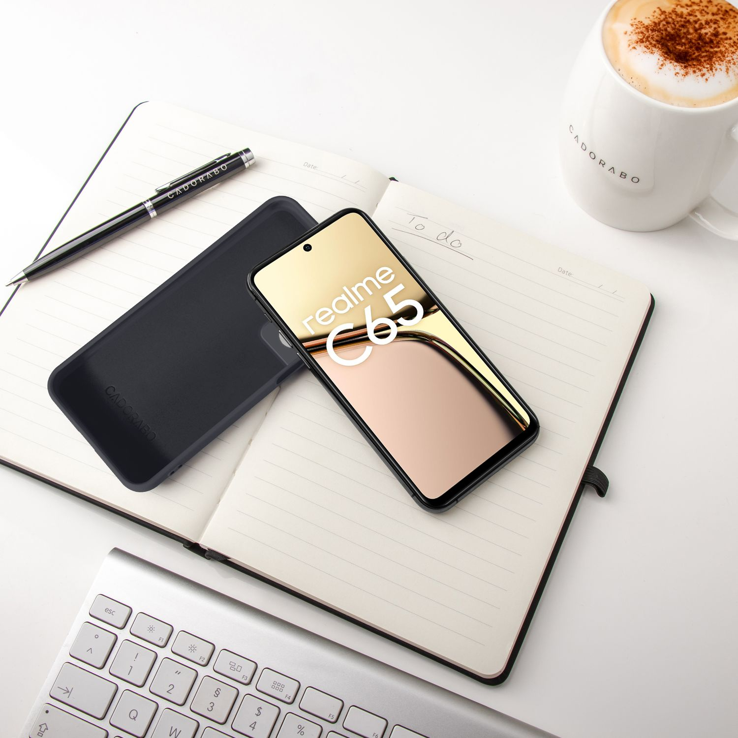 Ein Smartphone, Notizbuch, Stift, Tastatur und eine Kaffeetasse auf einem weißen Tisch.