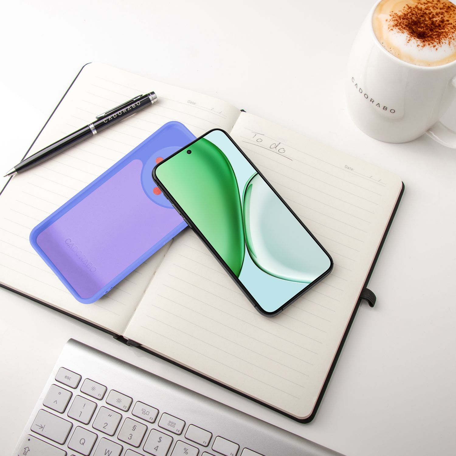 Ein Handy, ein lilafarbenes Etui, ein Notizbuch, Stift und Kaffee auf einem Schreibtisch.
